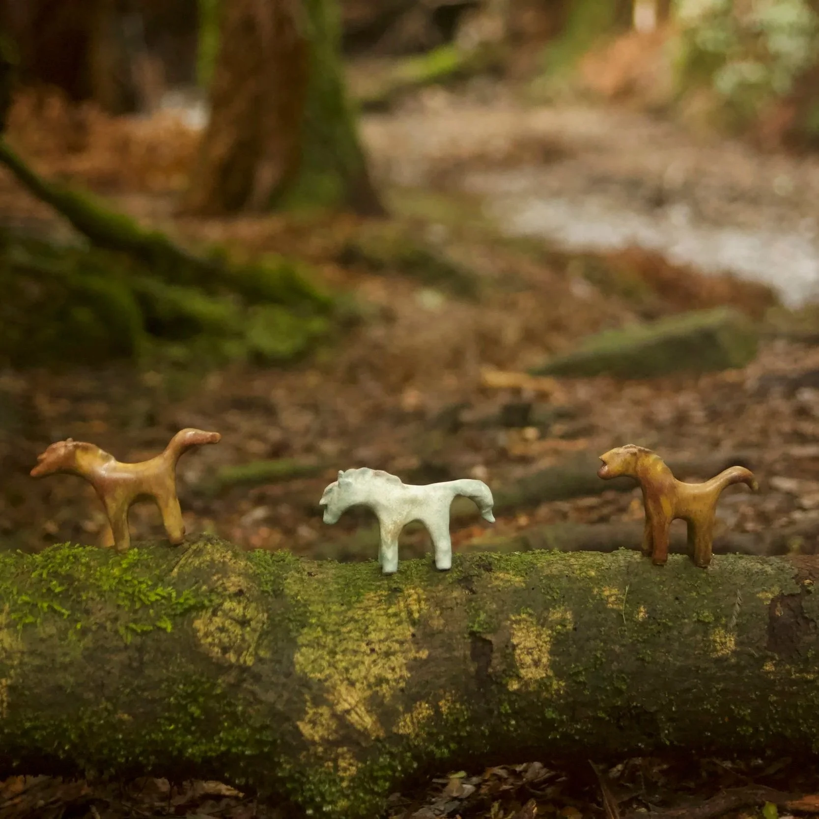 Three small toy horse figurines placed on a moss-covered log in a forested area with trees and a stream in the background.