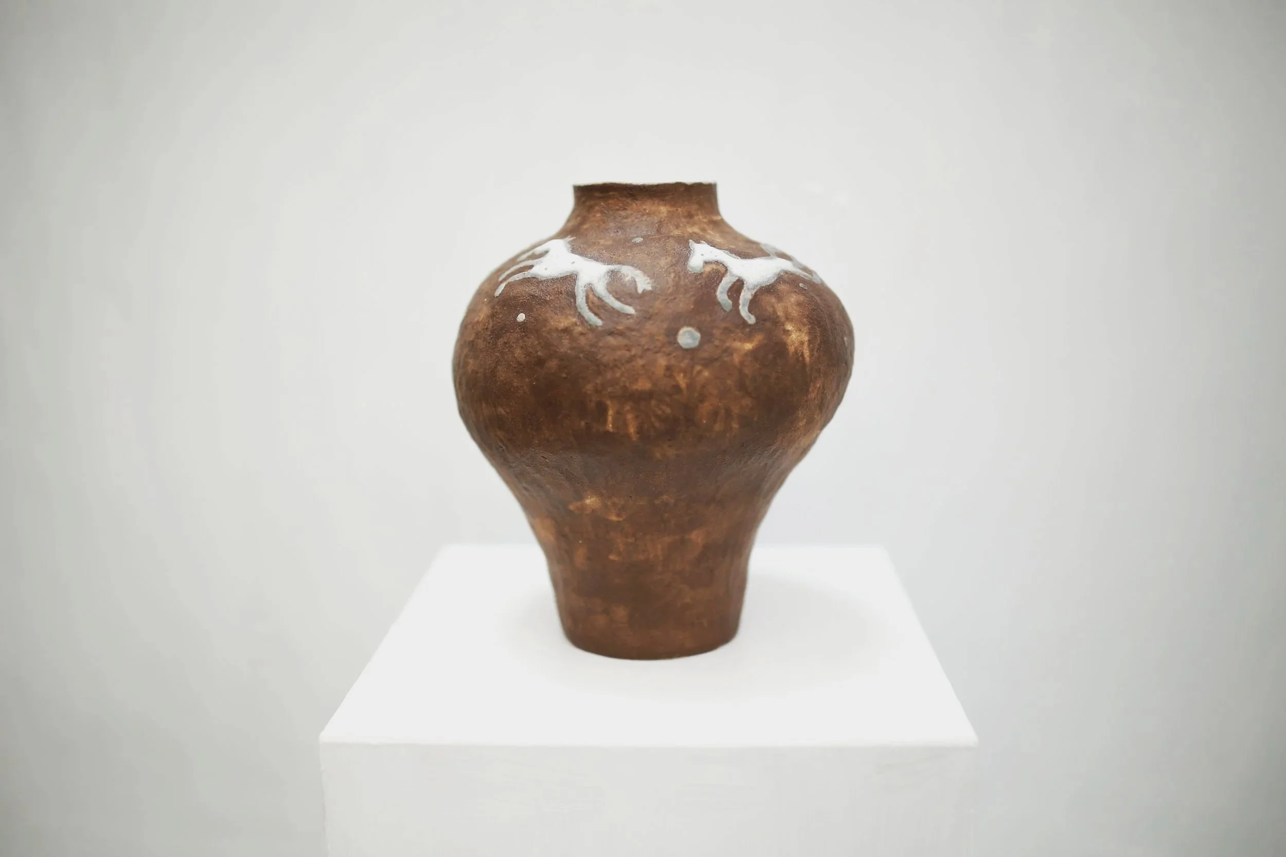A brown ceramic vase with white horse designs displayed on a white pedestal against a plain background.