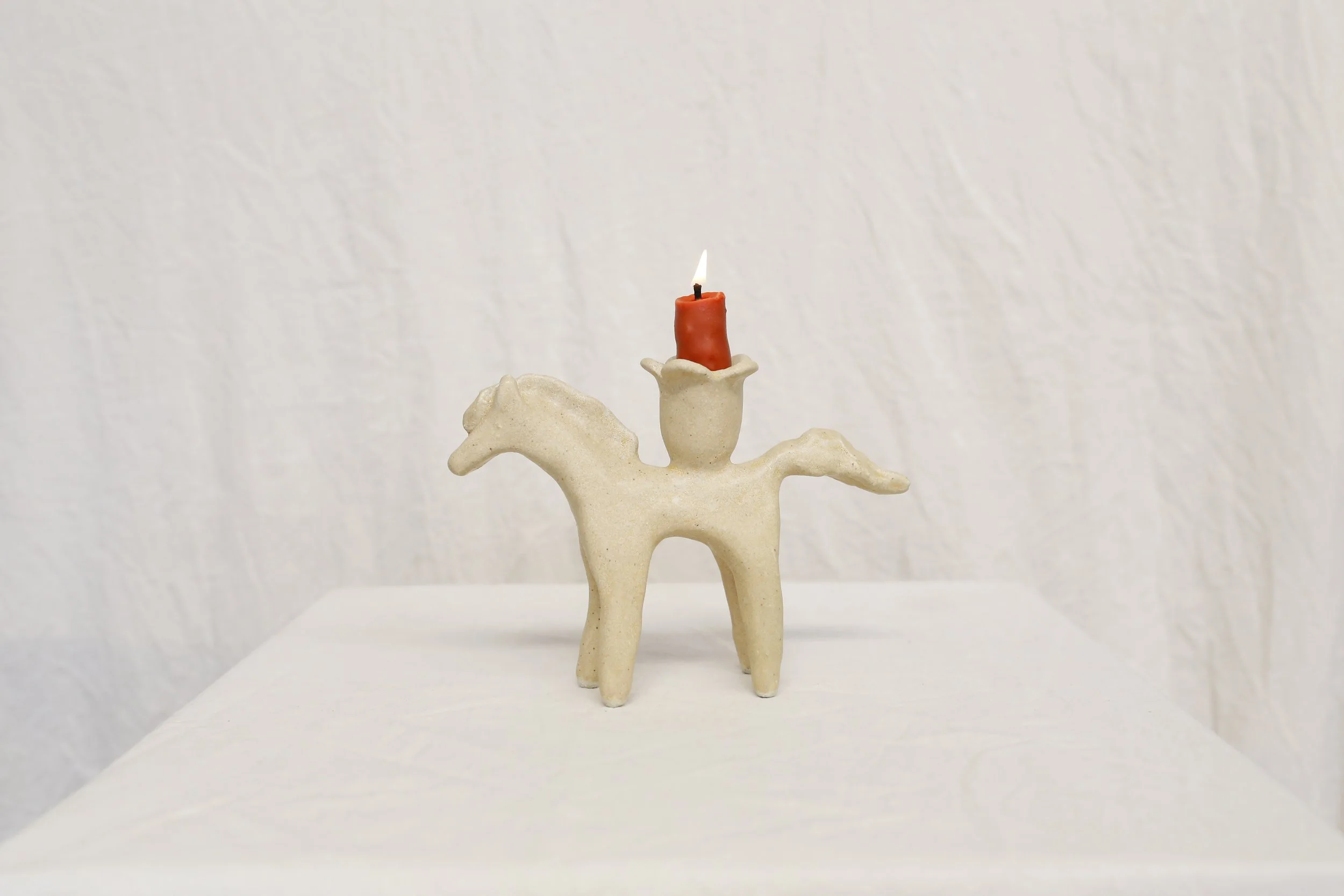Ceramic horse figurine with a lighted red candle on its back, placed on a white surface against a white textured background.