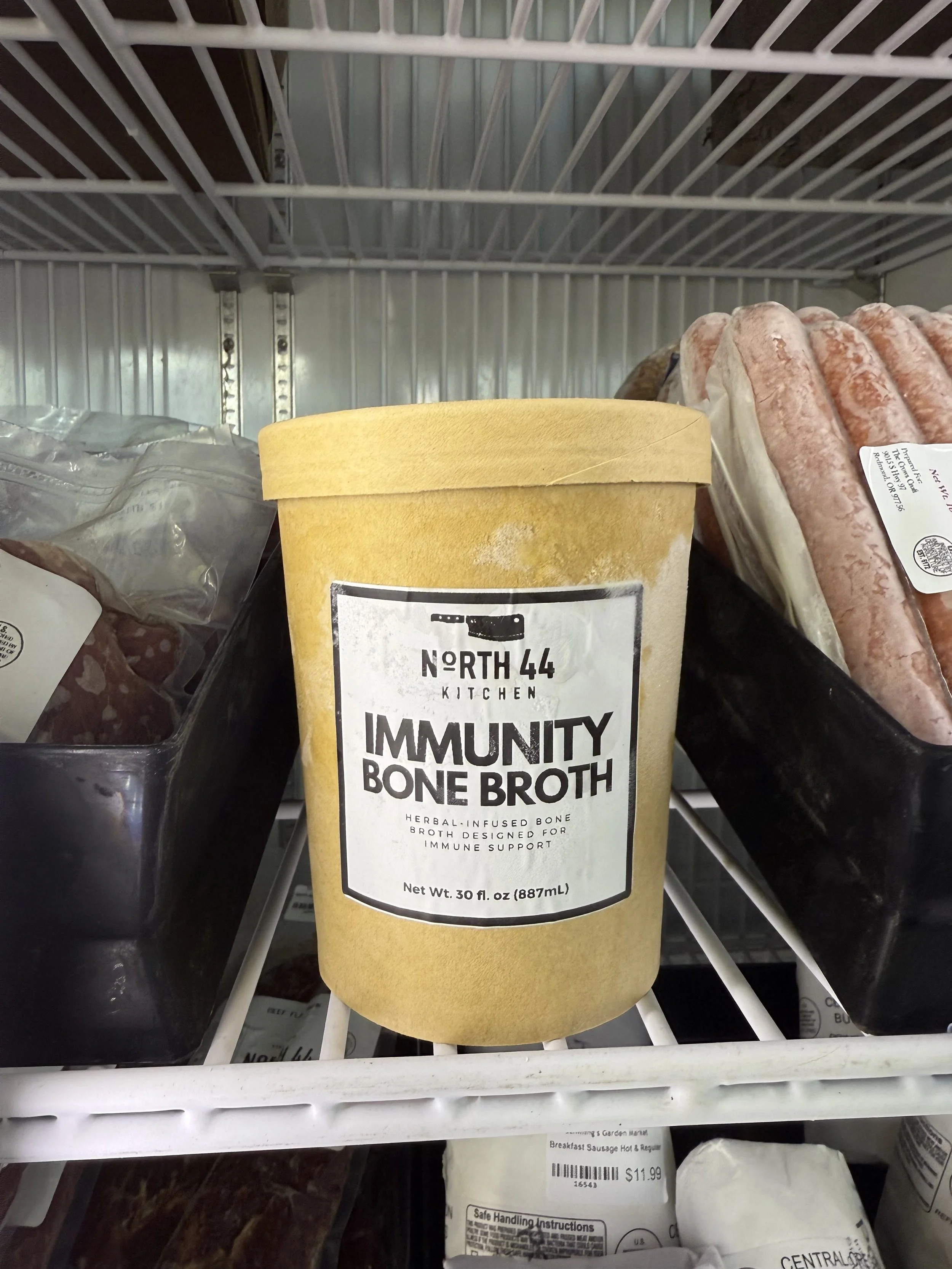 A yellow container of North 44 Kitchen Immunity Bone Broth on a refrigerator shelf, with raw sausages and other food items surrounding it.