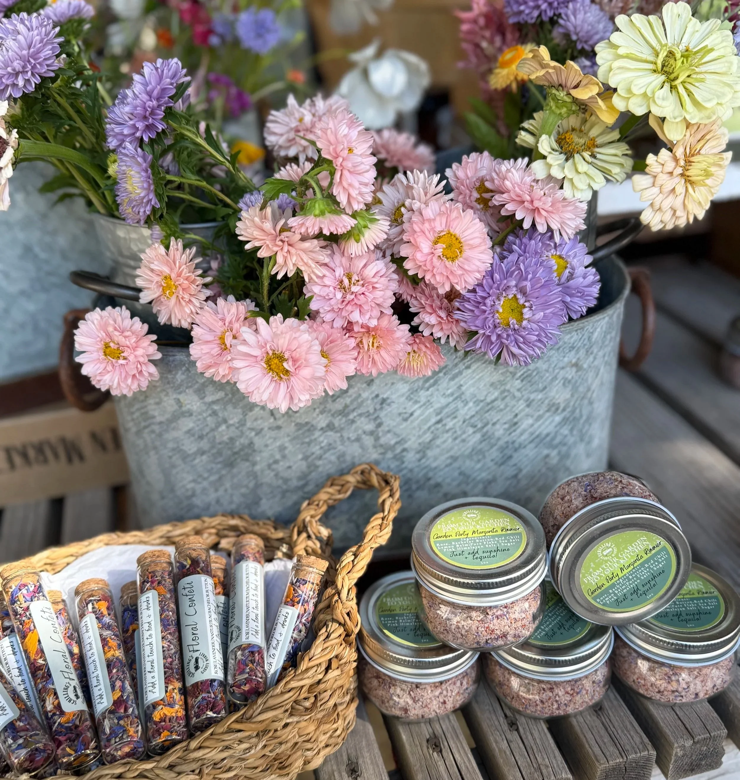 A metal container filled with pink, purple, and white flowers, placed on a wooden surface. Next to it, there are small jars labeled 'Garden Party Margarita Rimmer' and a basket holding plastic tubes filled with dried flowers, likely herbal or flower