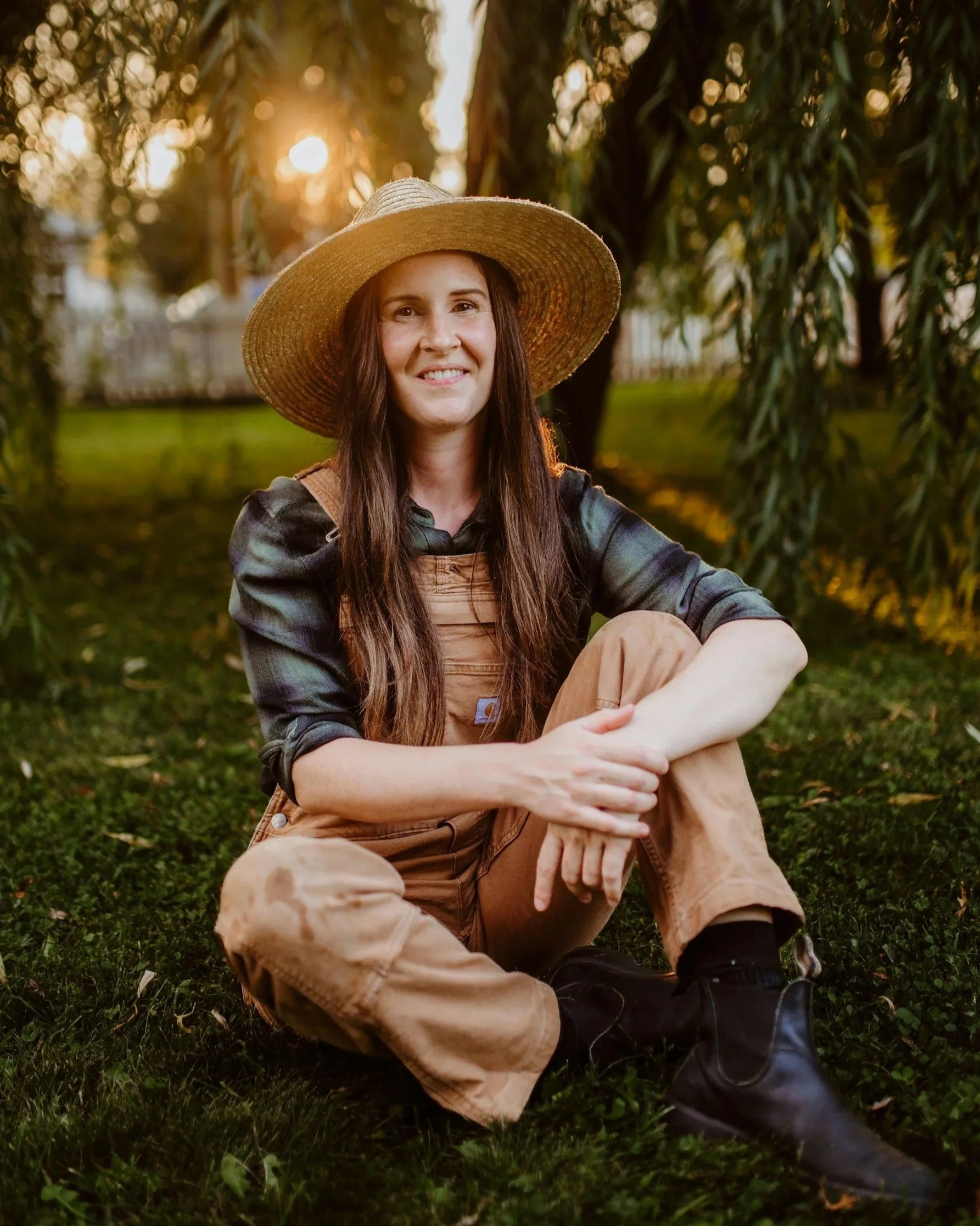 A woman sitting on the grass in a park at sunset, wearing a large straw hat, plaid shirt, overalls, and black boots, smiling at the camera.