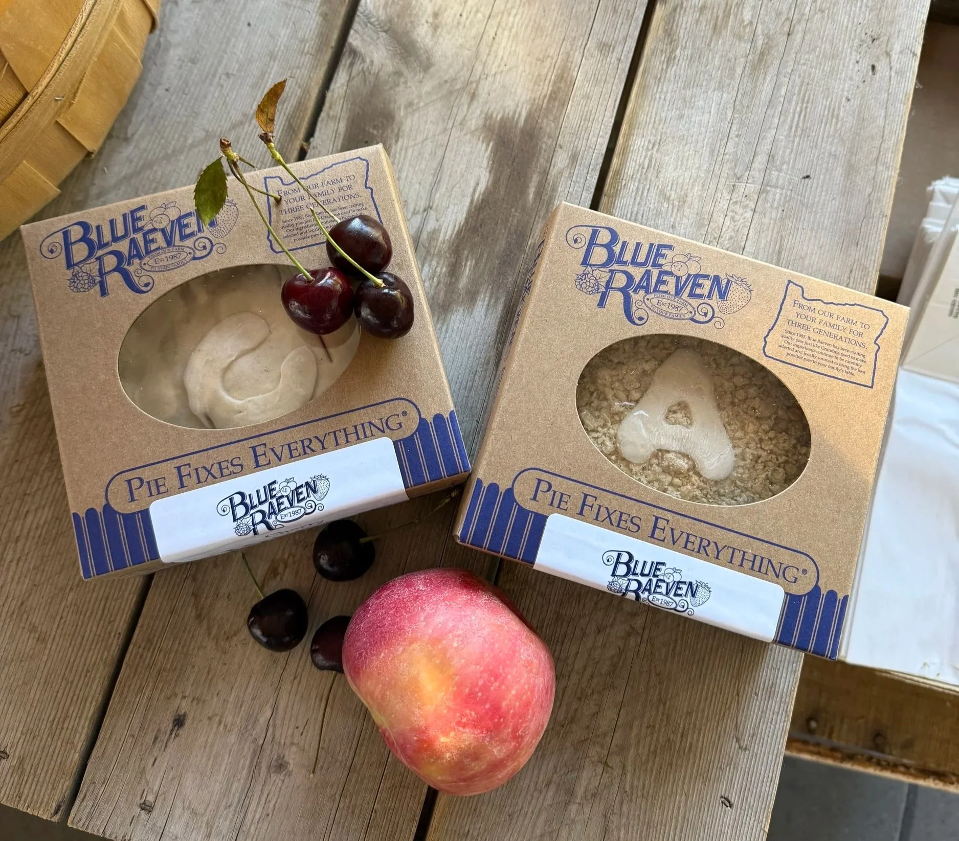 Two boxes of Blue Raeven pie fixings on a wooden table, one with cherries on top and the other with crumbled pie crust, a peach, and some cherries beside them.
