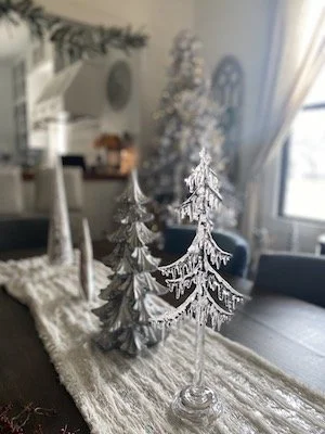 A decorative display featuring silver Christmas tree ornaments placed on a textured table runner in a dining room. The background includes a window with curtains and a hint of a festive ambience.