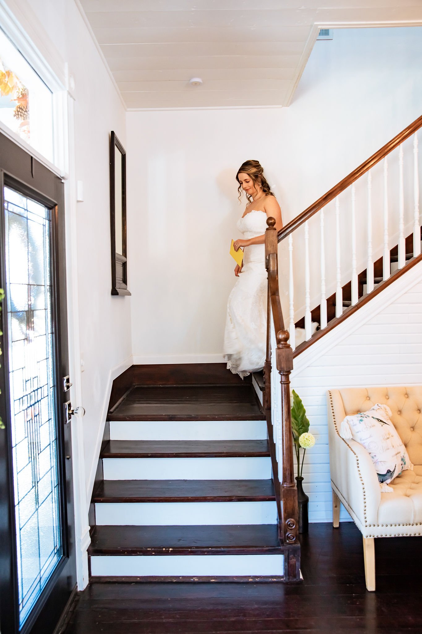 Bride in a white dress descending wooden stairs in a well-lit room with a beige armchair and a decorative plant on the side.