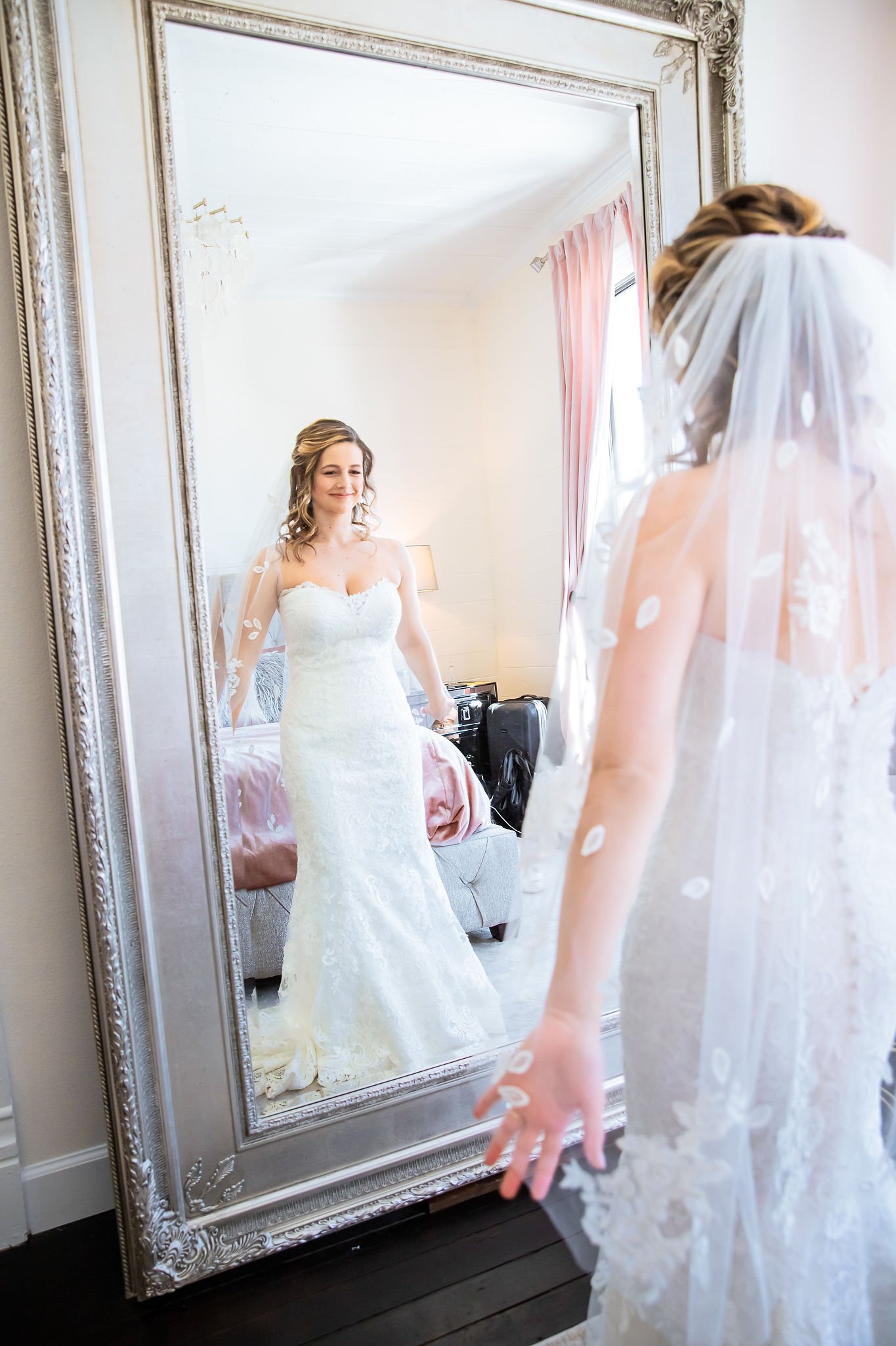 Bride in a lace wedding dress and veil looking at herself in a large ornate mirror in a room with pink curtains.