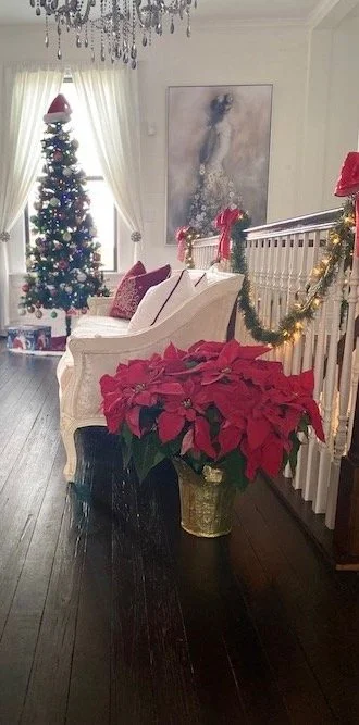 A festive living room decorated for Christmas with a beautifully adorned Christmas tree near a window. The room features white curtains, a white bench with red cushions, a potted poinsettia plant in front, and a stair railing wrapped in greenery and lights. A chandelier hangs from the ceiling.