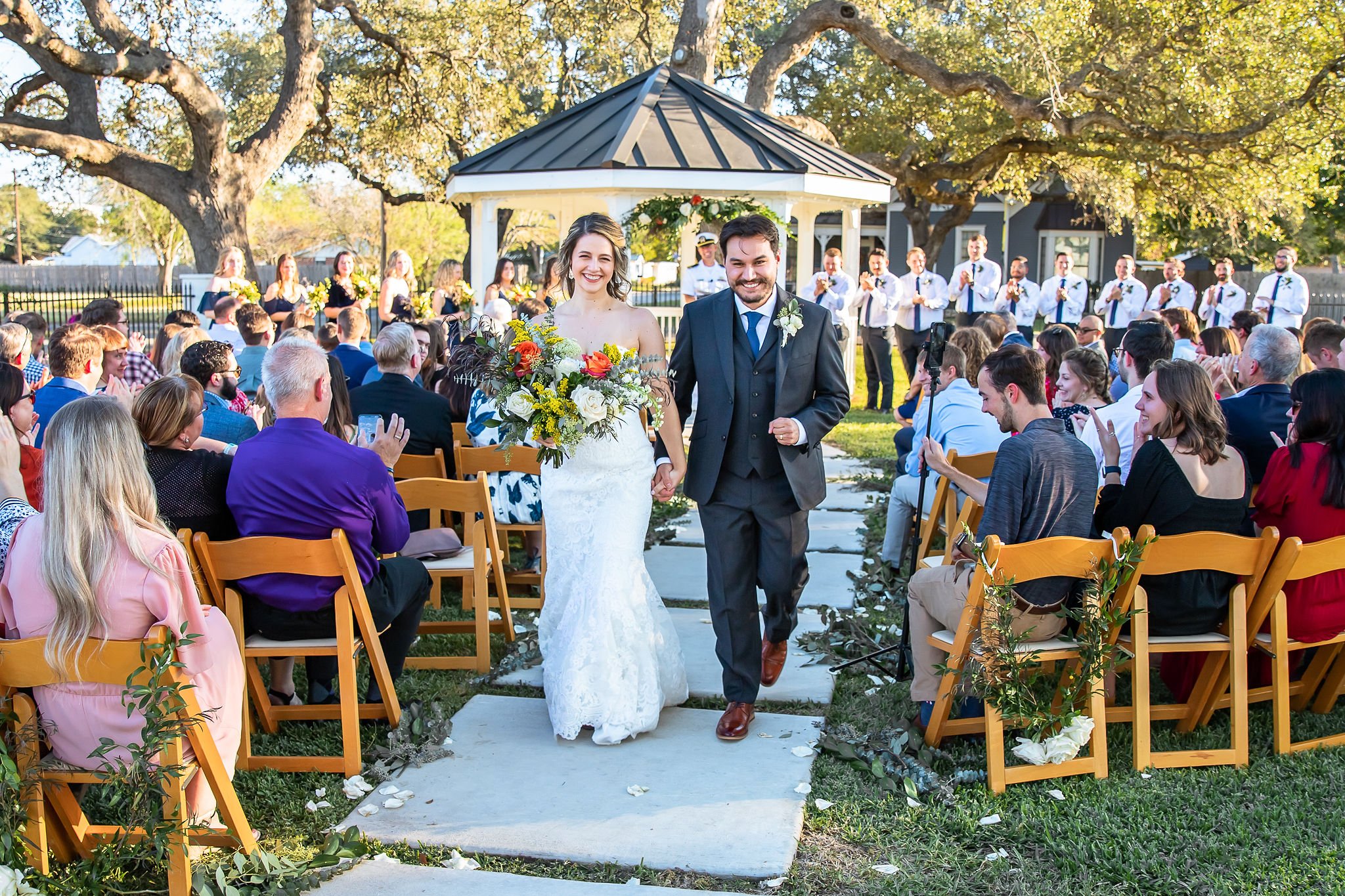 A bride and groom walking down the aisle outdoors, smiling and holding hands, with a gazebo in the background and guests applauding on both sides.