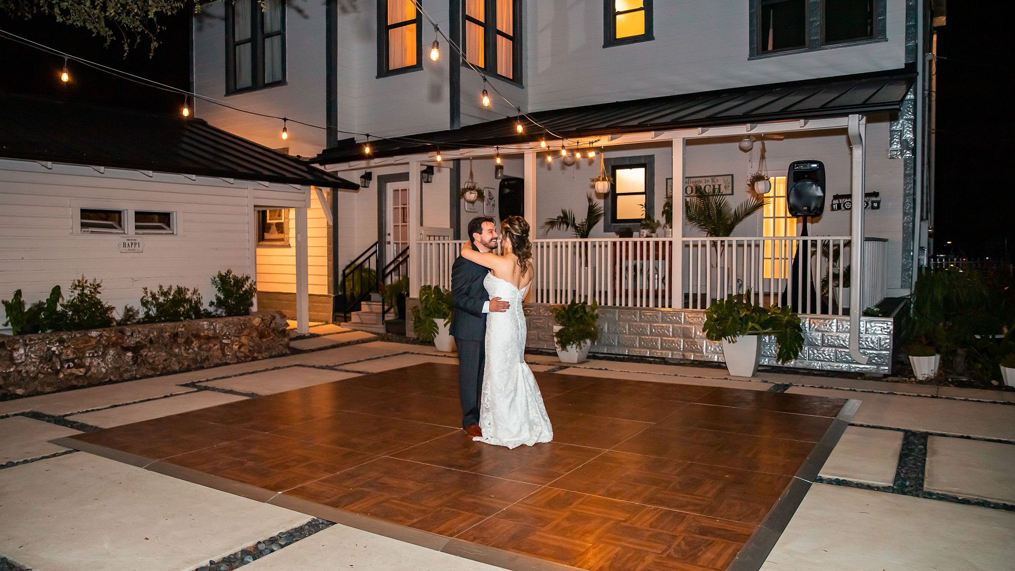 Bride and groom dancing outdoors at night on a wooden dance floor with string lights above, in front of a white house with a porch.