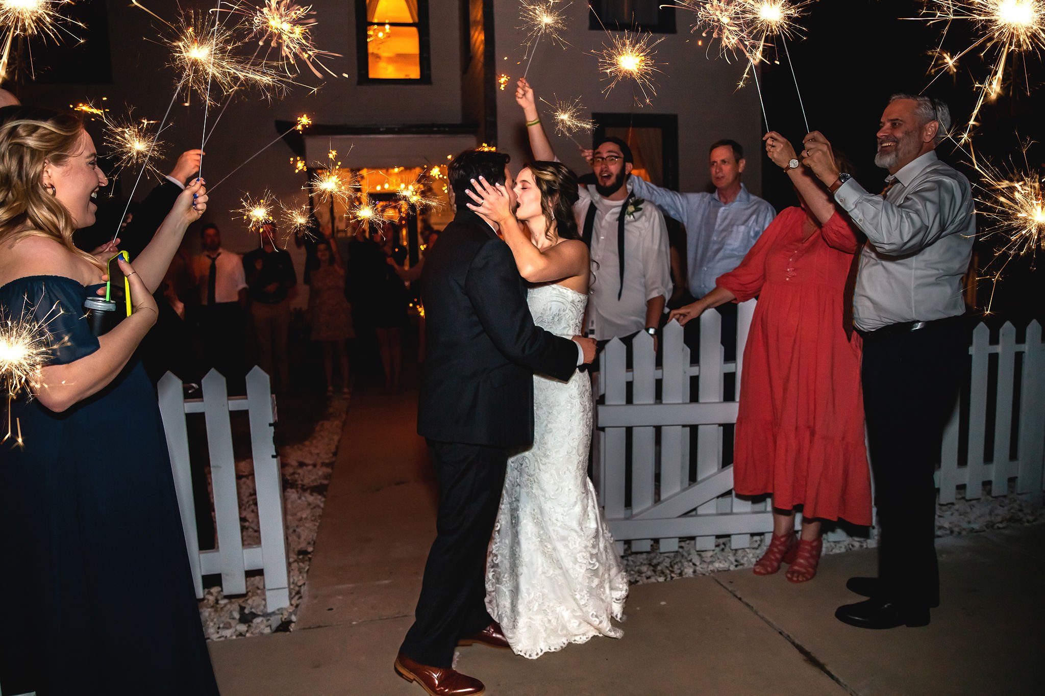 Couple in wedding attire kissing at a night celebration with people holding sparklers around them.