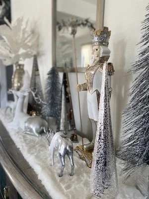 Christmas-themed mantel display featuring silver and white decorations including a nutcracker, reindeer figurines, and artificial trees.