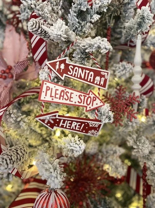 Snow-covered Christmas tree with ribbons and ornaments, featuring a "Santa Please Stop Here!" sign.