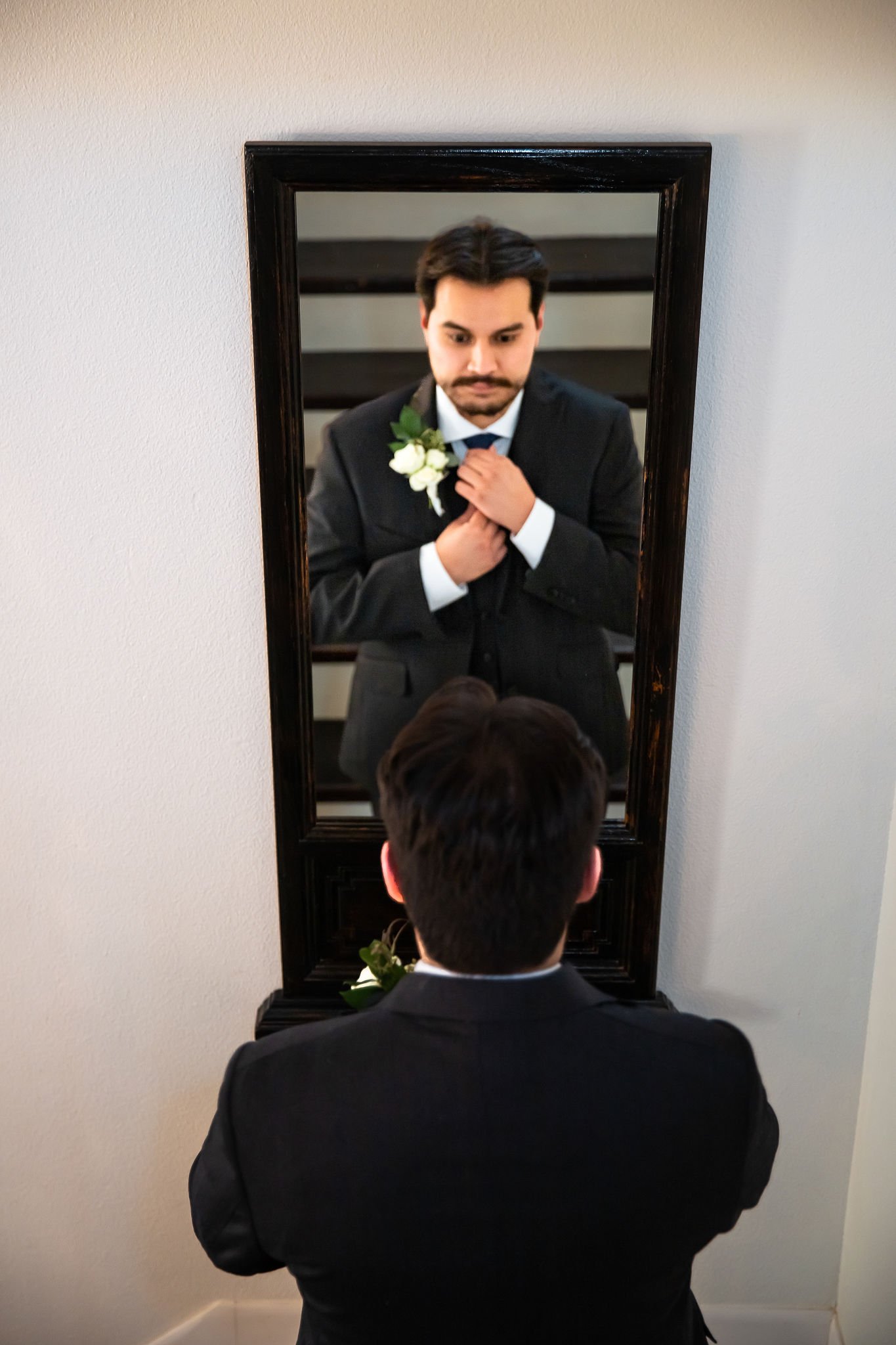 Groom adjusting tie in front of mirror, wearing a suit with a white boutonniere.