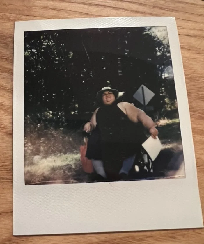 An image of a polaroid picture of Kennedy in her power wheelchair wearing a black cotton sun dress and sun hat. She is on the perimeter of a sunny wooded property and holding papers.
