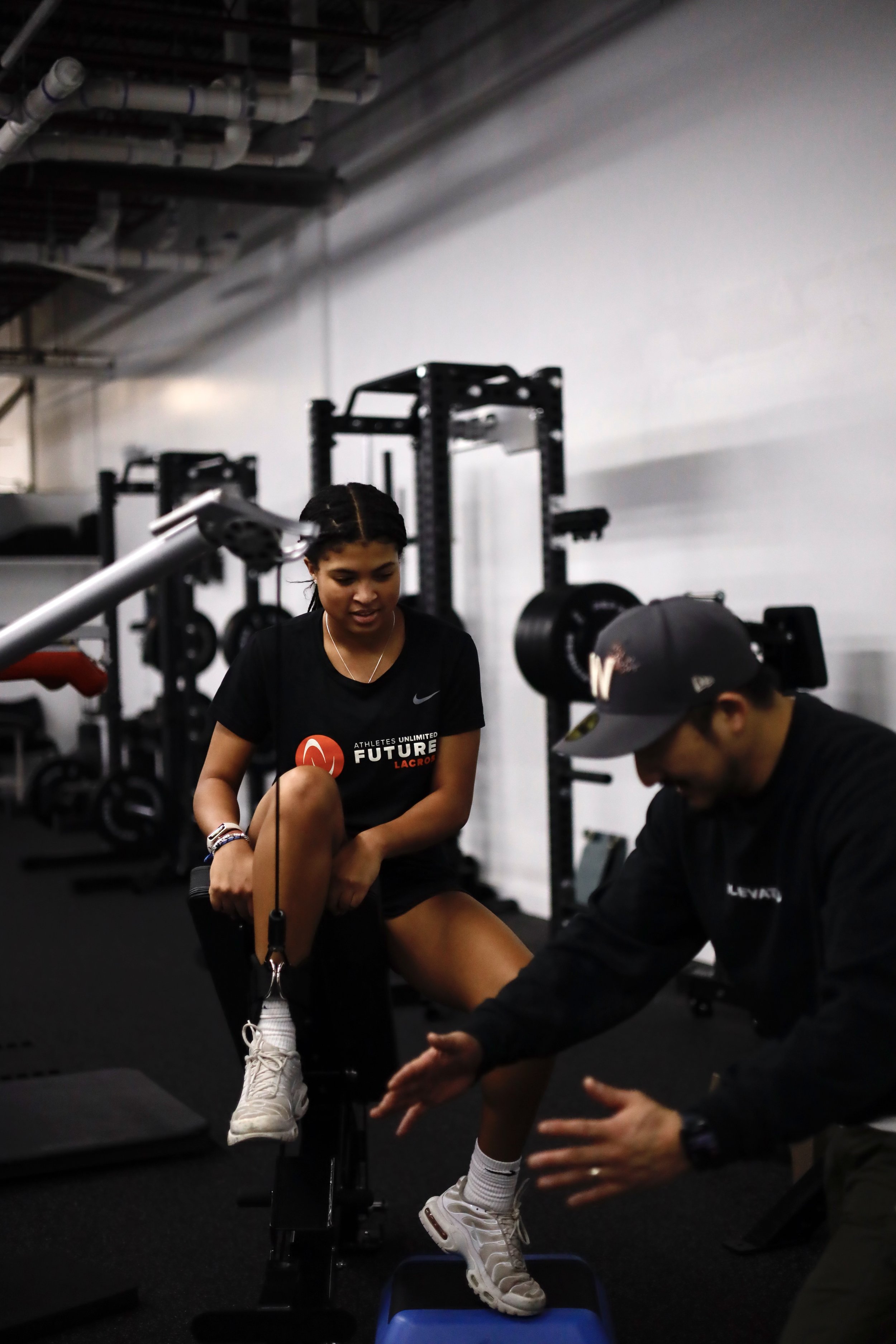 Person in a gym setting using exercise equipment, wearing a black t-shirt and white sneakers, alongside another person who appears to be a trainer.