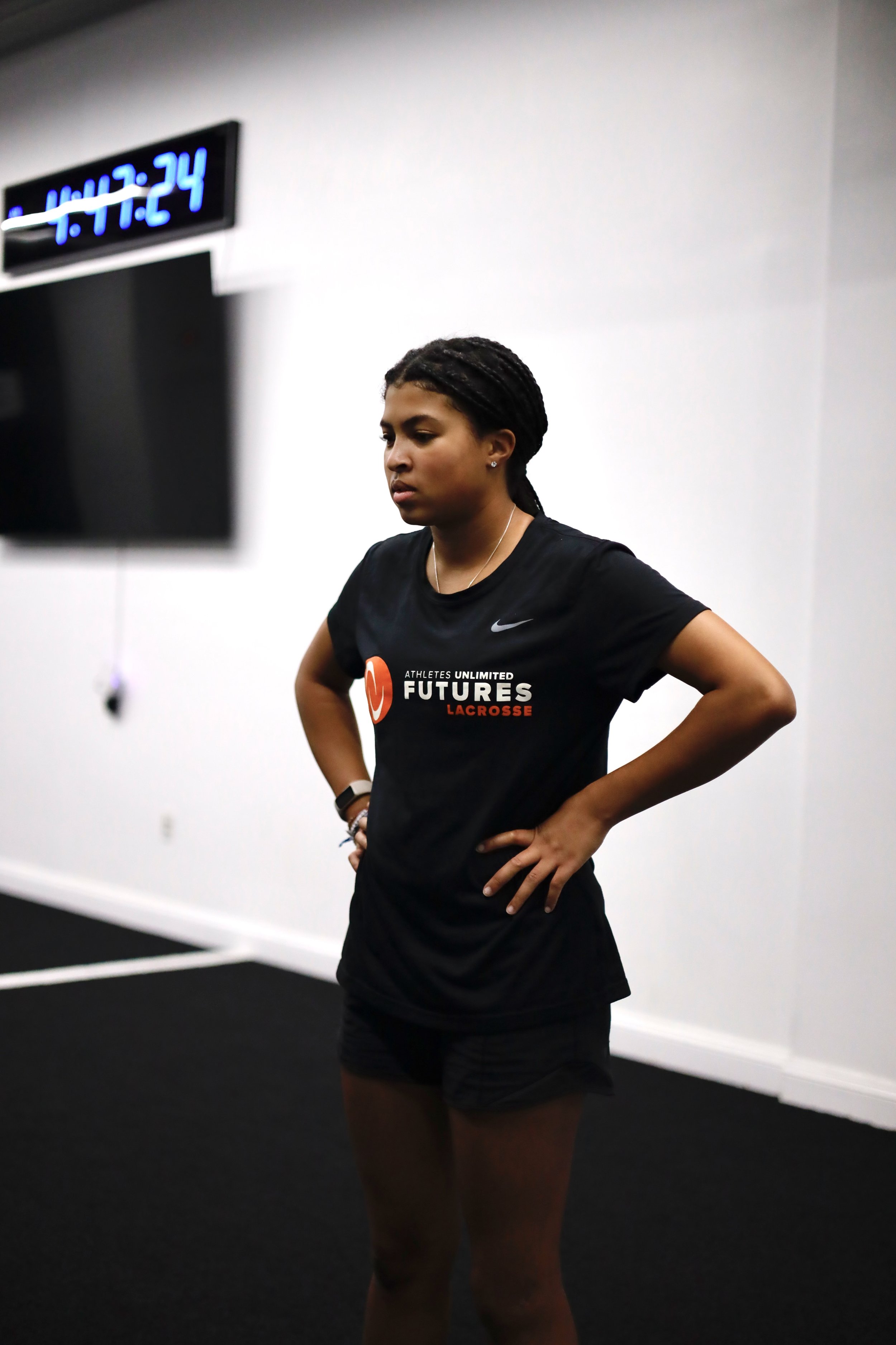 Person wearing a "Futures Lacrosse" t-shirt standing with hands on hips in a gym.
