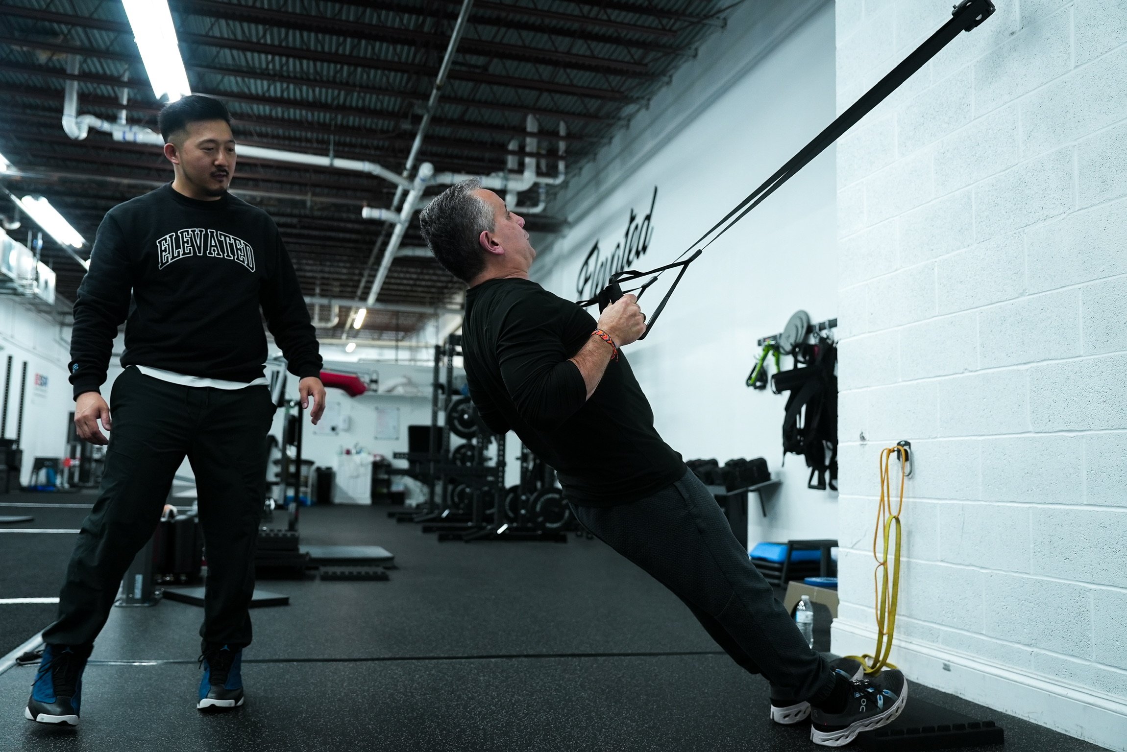 A man is performing a resistance exercise using a TRX suspension trainer in a gym, guided by a trainer standing nearby.