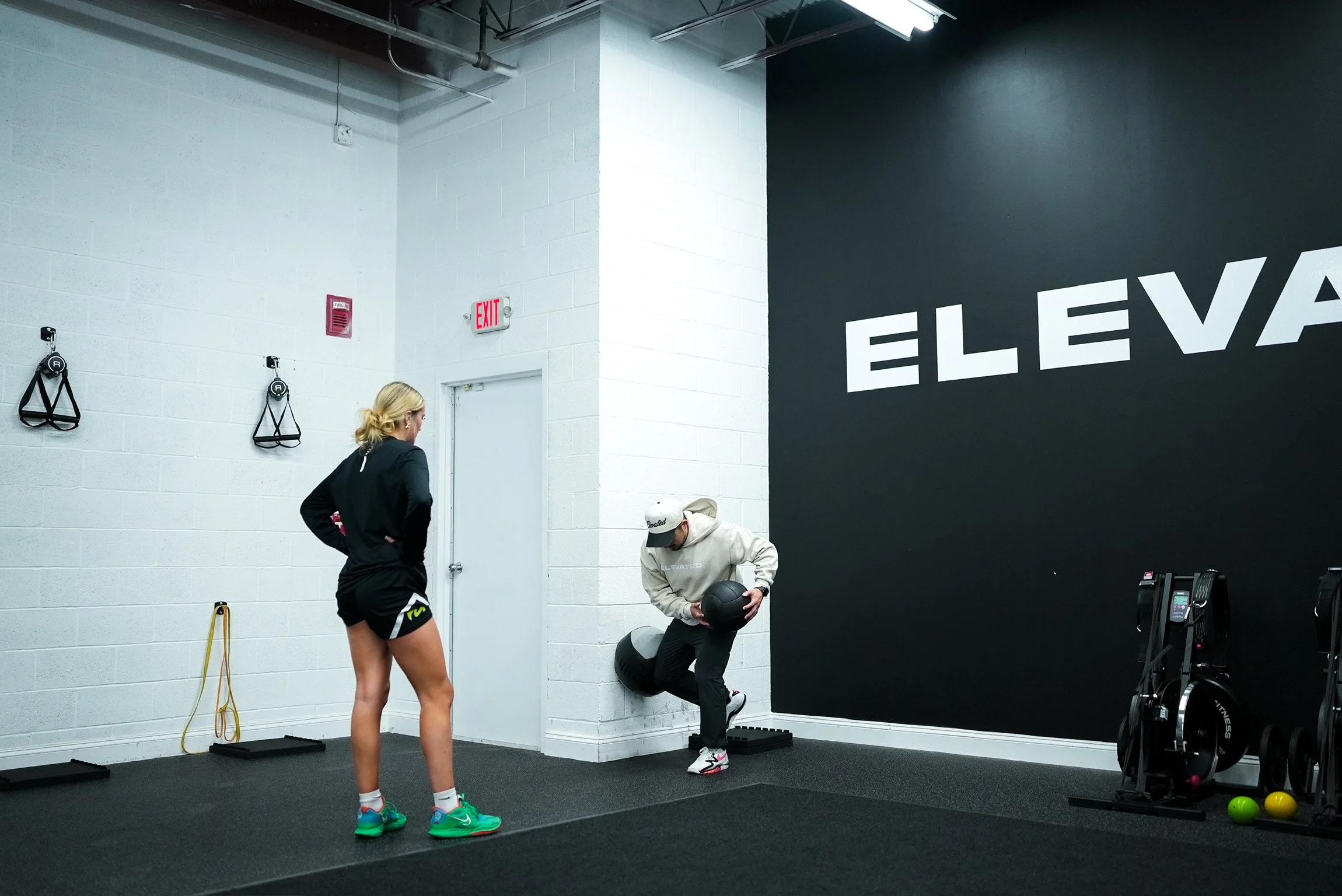 A woman and a man exercising in a gym. The woman is standing with hands on hips, wearing black workout clothes and colorful sneakers. The man is holding a medicine ball, wearing a white hoodie, black pants, and a white cap. The gym has white and black walls with partial text 'ELEV' visible. Fitness equipment like resistance bands, a foam roller, and medicine balls are on the floor.