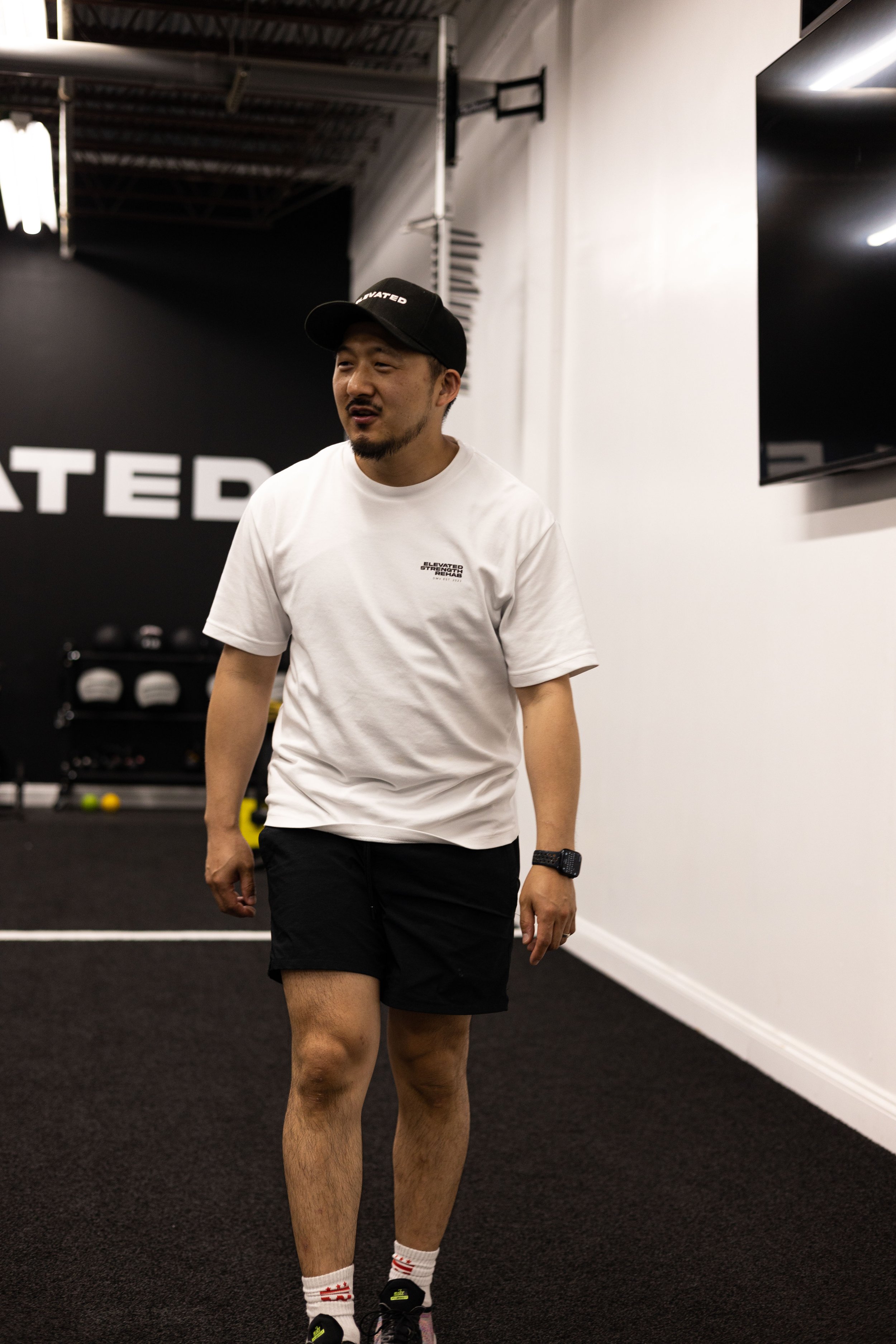 Man wearing a white shirt and black shorts in a gym setting, with exercise equipment in the background.