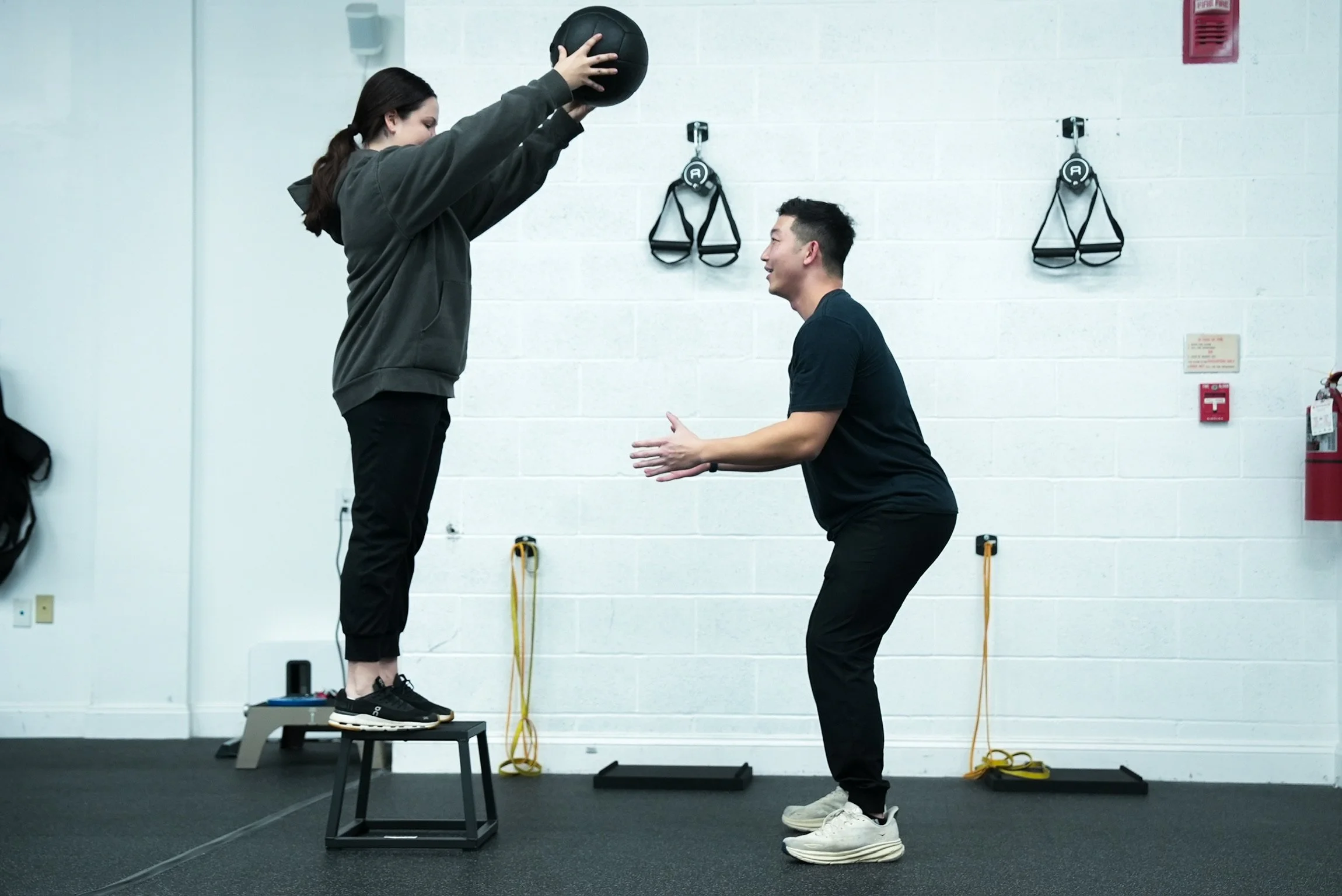 A woman standing on a workout step holding a black medicine ball above her head, with a trainer in front of her kneeling and clapping in encouragement in a gym.
