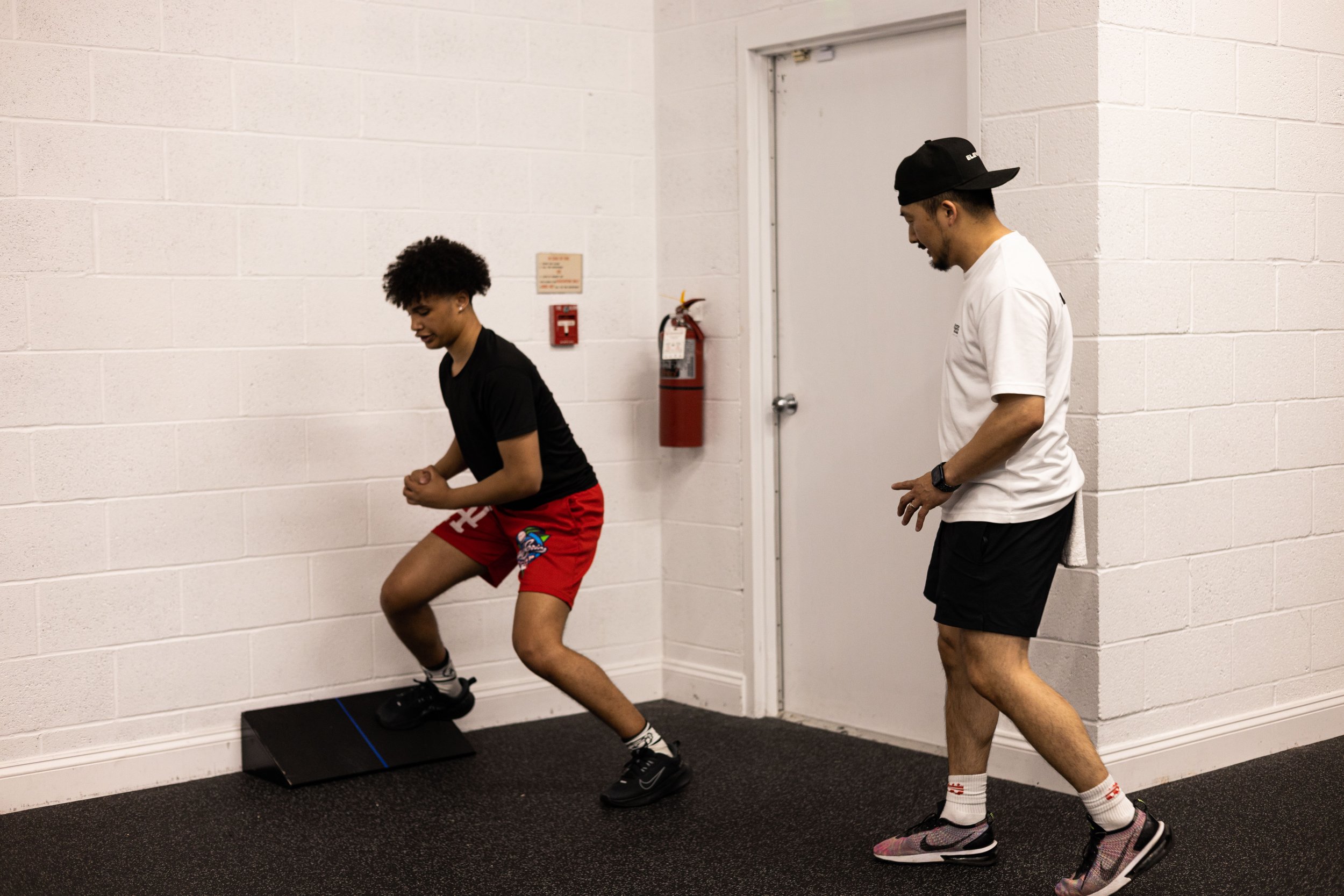 Two people in a gym setting, one performing a squat exercise with a slant board against a wall, while the other observes and gives guidance.