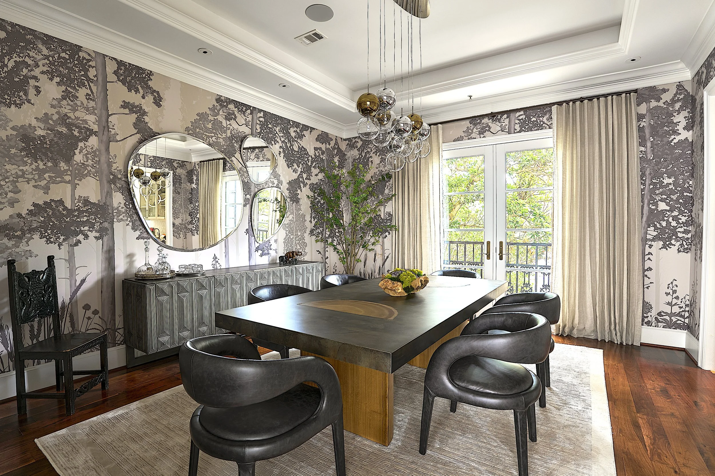 A dining room with a large dark wood table surrounded by six black leather chairs. The room features wallpaper with a tree and forest design, a rustic sideboard with decorative items, circular mirrors on the wall, and a modern chandelier with multiple glass orbs hanging from the ceiling. Large windows with cream curtains let in natural light.