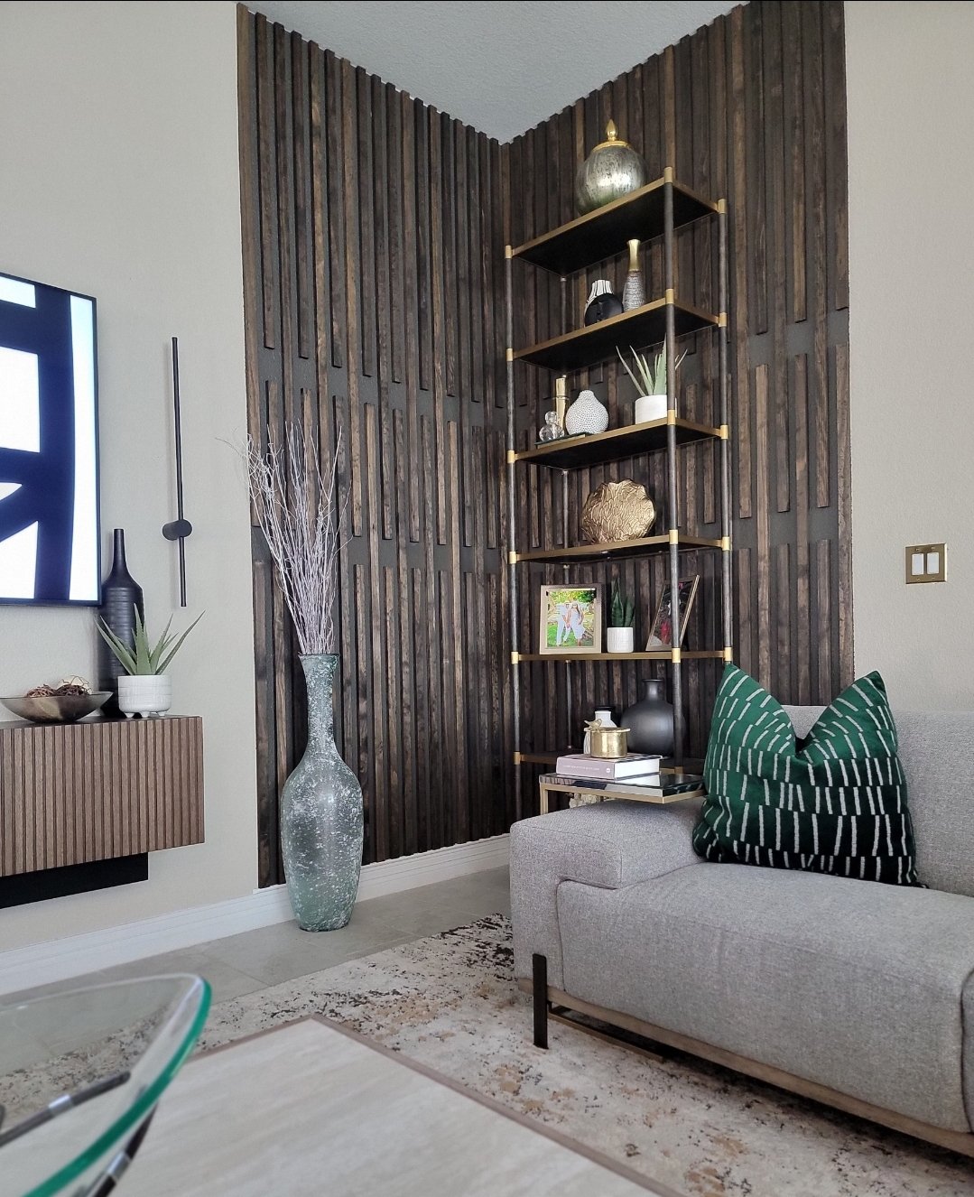 Living room corner with a beige sofa, striped green and black pillow, black and gold shelving unit with decorative vases and plants, dark wooden accent wall, large glass vase with decorative twigs, wall-mounted TV, and a small wall-mounted wooden cabinet with a potted plant.