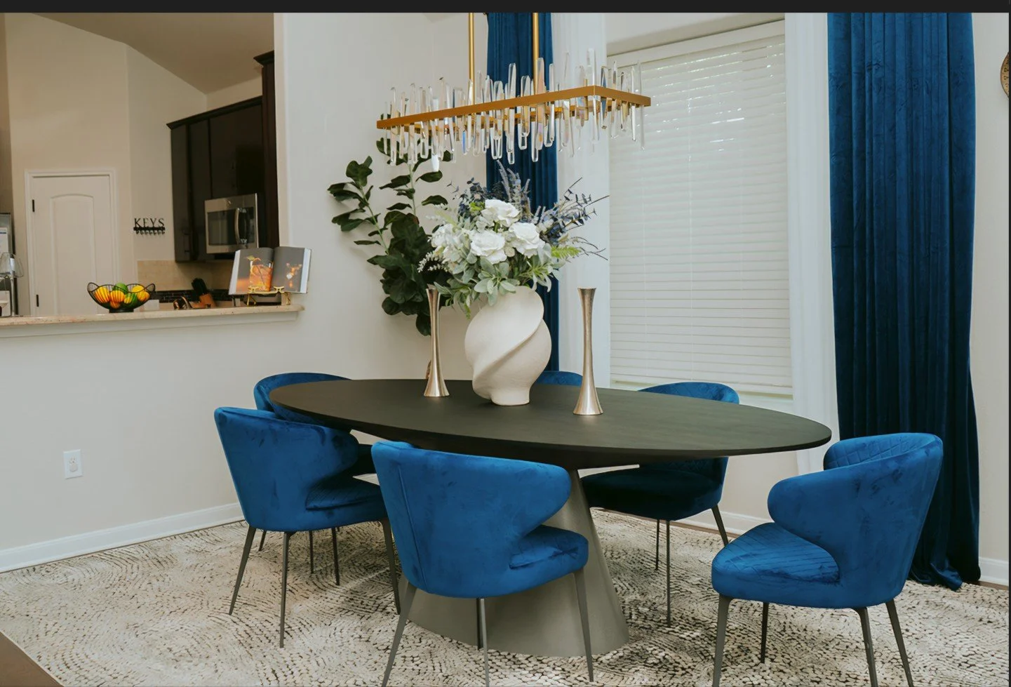Modern dining room with a black oval table, six blue velvet chairs, a large floral centerpiece, two gold vases, a contemporary chandelier, and blue curtains in front of white blinds.