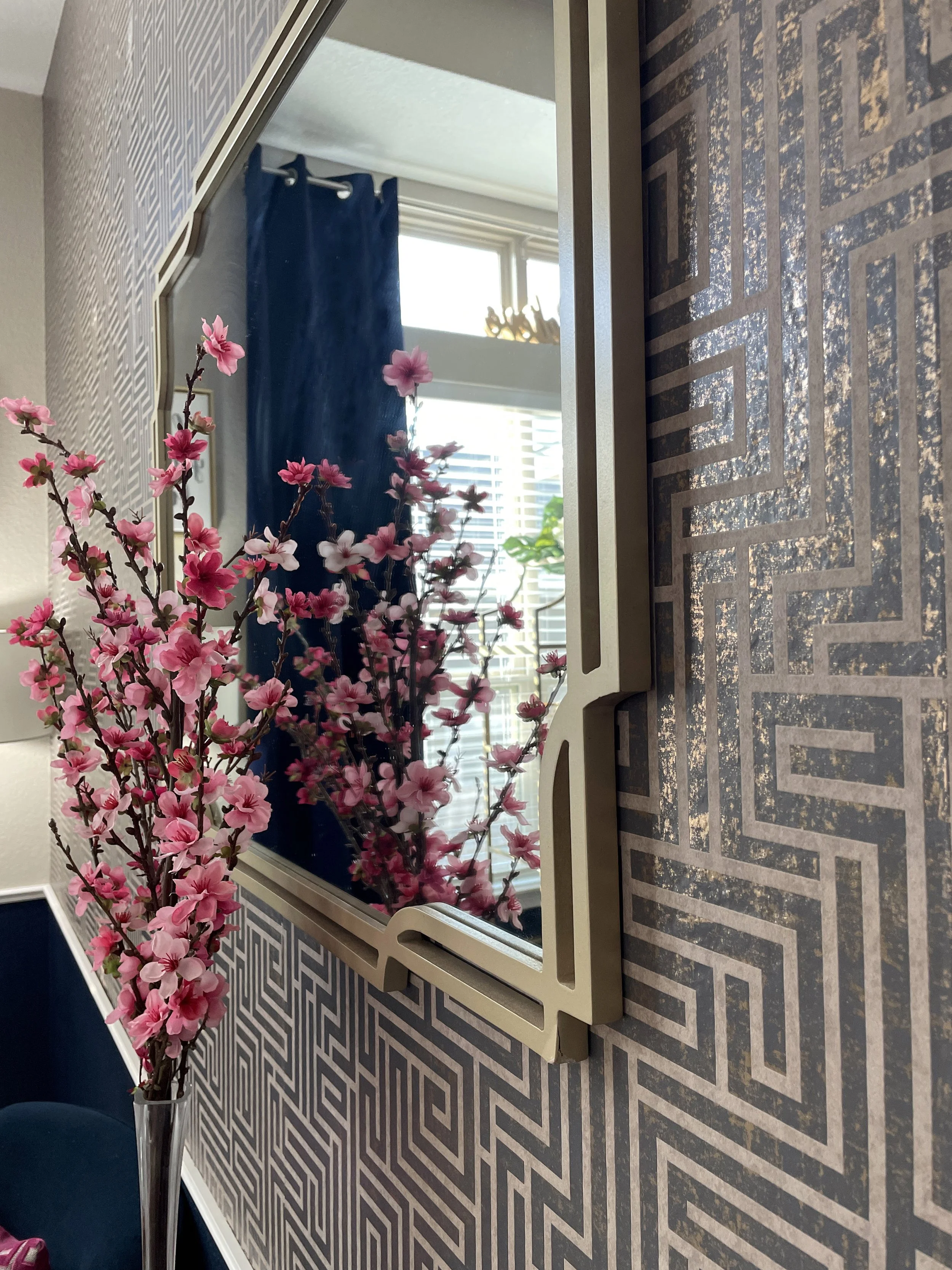 A close-up view of a decorative mirror with a marble-like patterned frame, placed on a patterned surface, with a vase of pink flowering branches reflected in the mirror, a window with closed blinds, and blue curtains in the background.