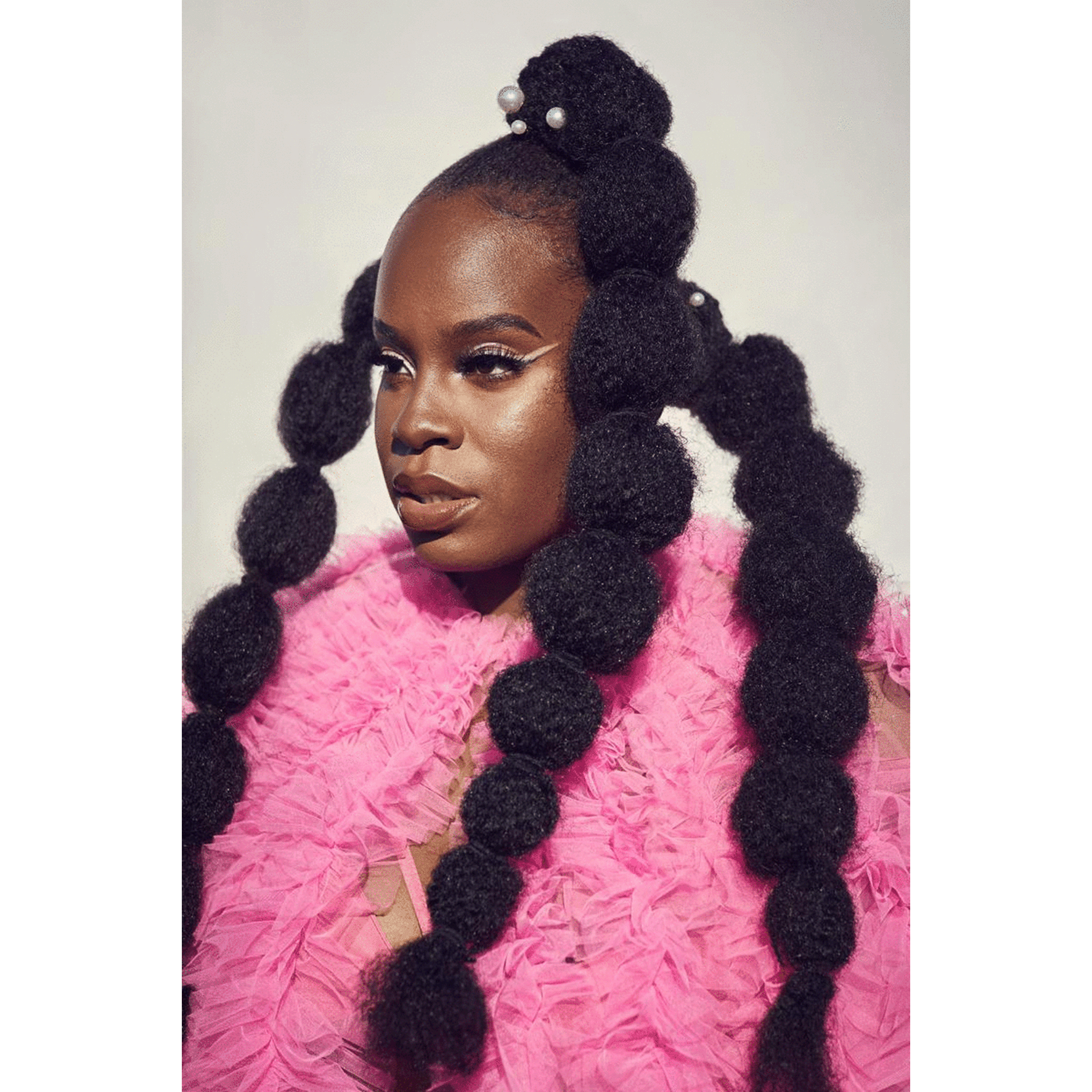 Stop motion gif in Dubai. A woman with dark skin and an elaborate hairstyle featuring large, textured puffs decorated with pearls, wearing a pink feathered garment against a neutral background.