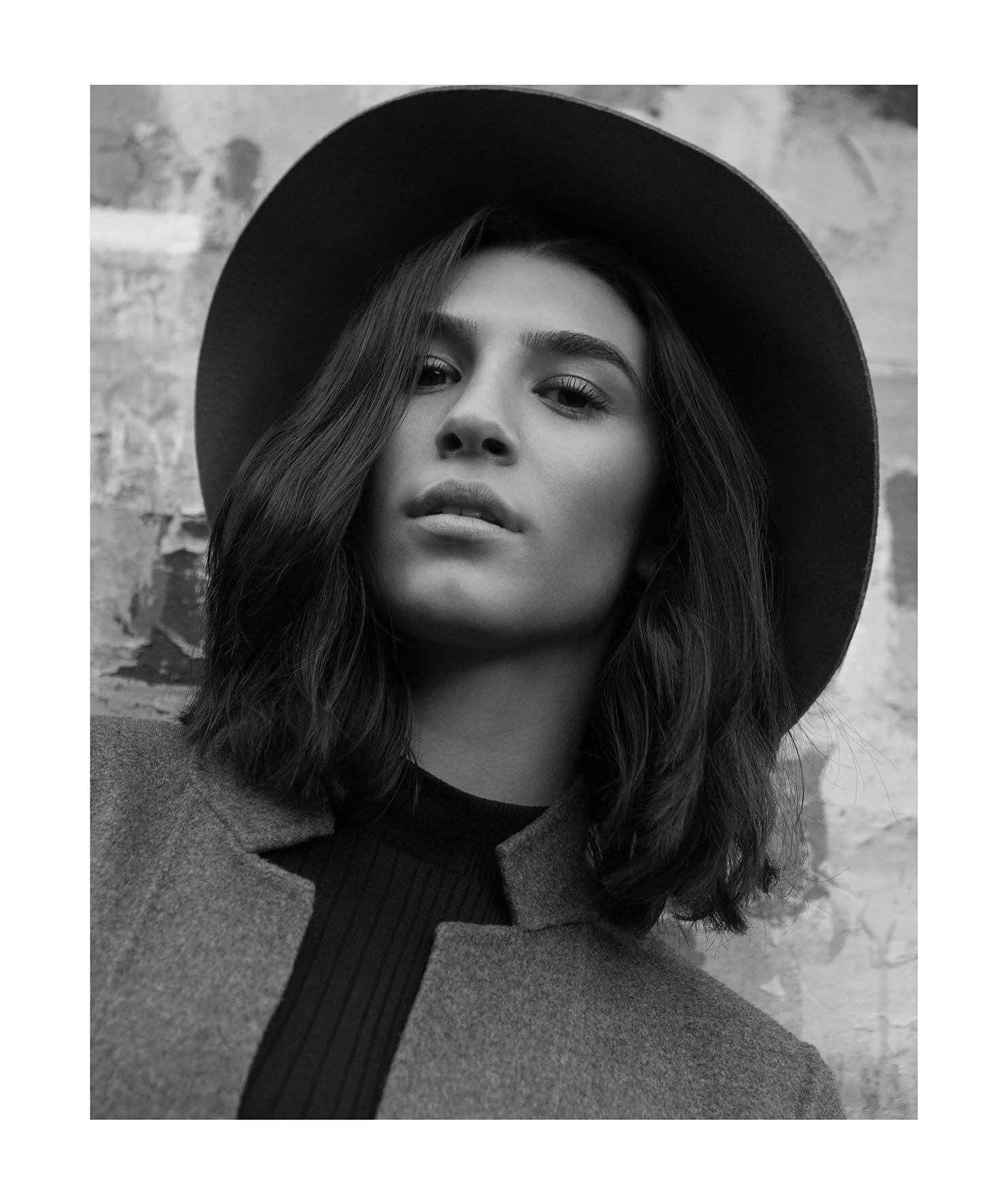 Editorial photography in Dubai. Black and white portrait of a woman with wavy hair wearing a wide-brimmed hat and a jacket, standing against a textured wall.