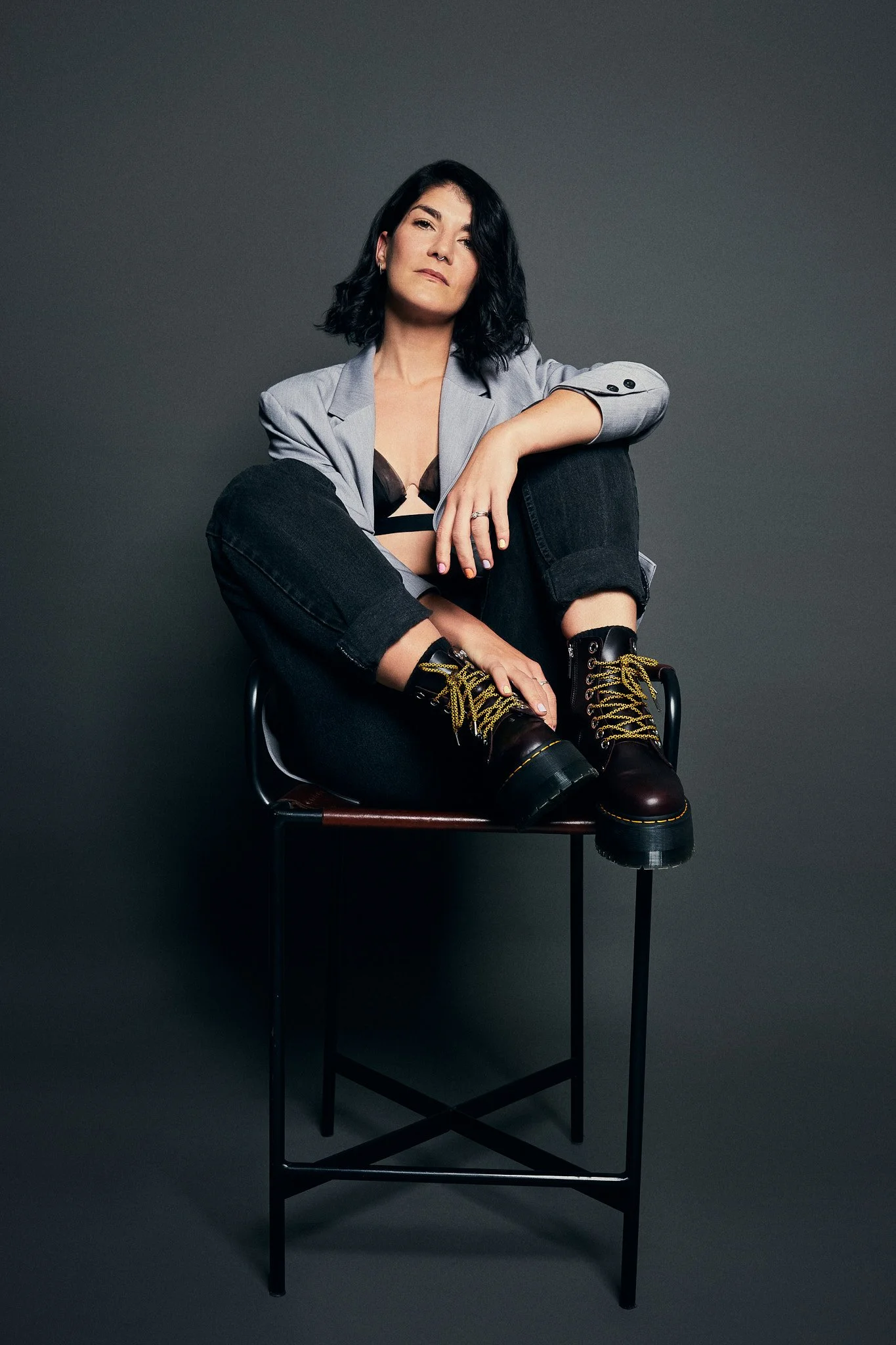 Portrait photographer in Dubai. A woman with black hair, wearing a gray blazer, black jeans, and black boots with yellow laces, is sitting on a chair against a plain dark gray background.