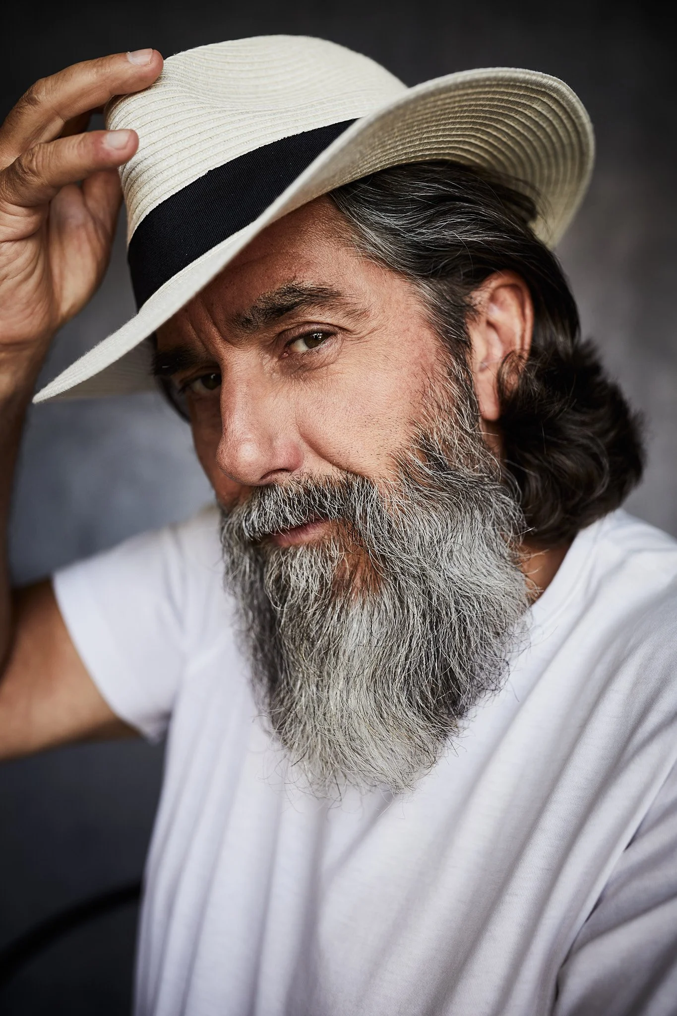 Actor Headshots portraits in Dubai. A middle-aged man with a gray beard, wearing a white t-shirt, holding a straw hat with a black band, and looking at the camera.