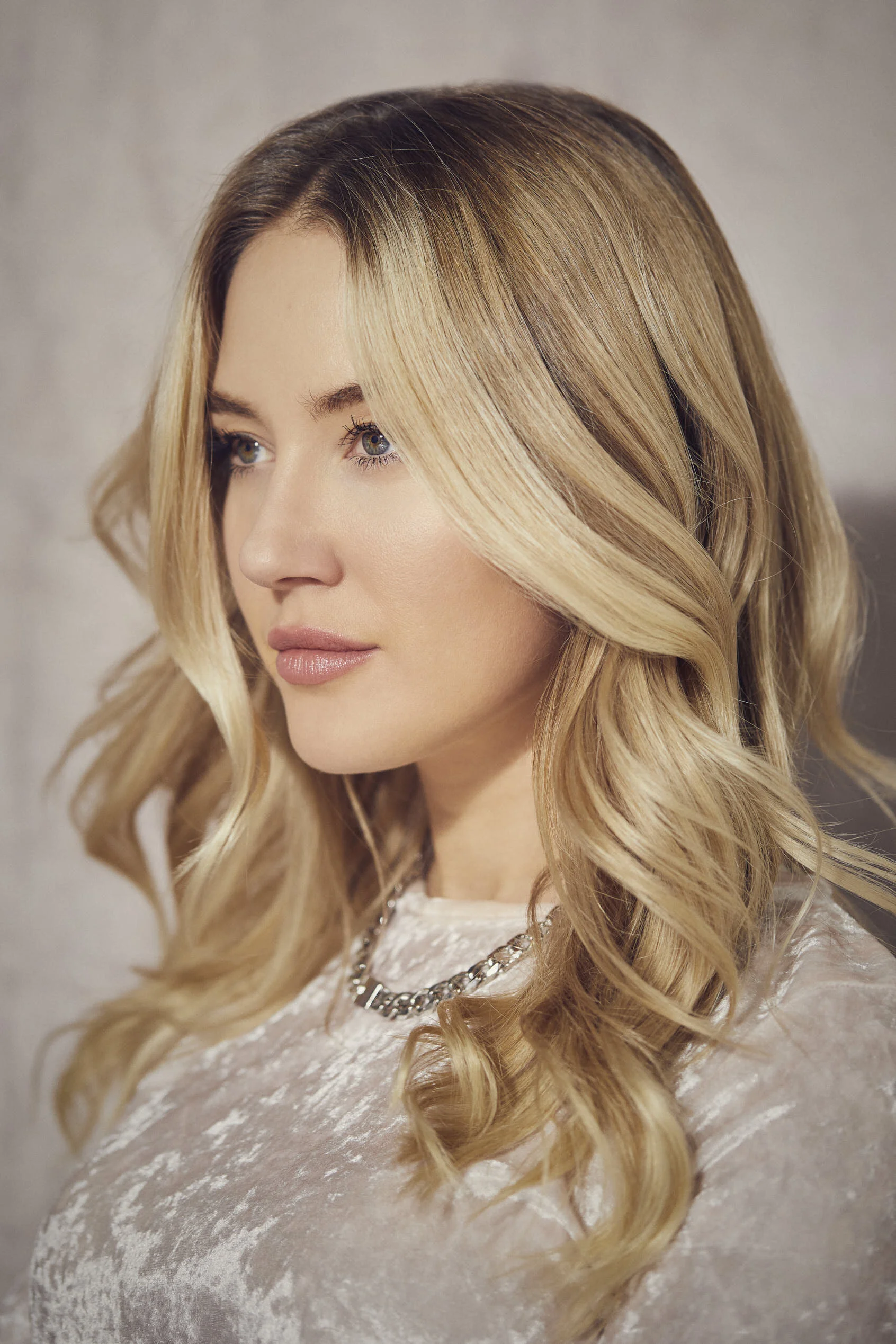 Actress headshot photography in Dubai. A woman with long, wavy blonde hair looking to the side, wearing a light-colored top and a silver chain necklace.
