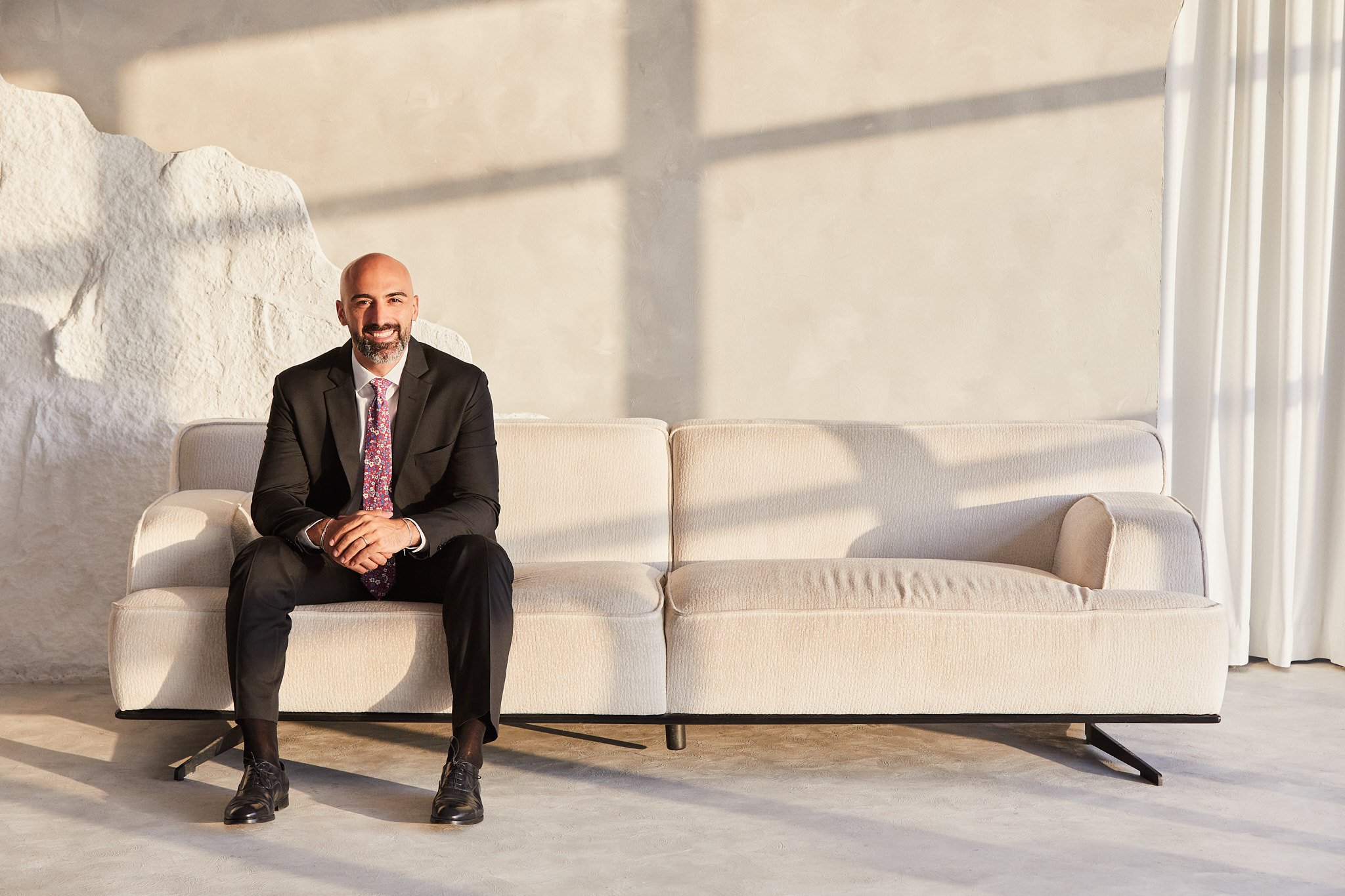 Portrait photographer in Dubai. A man with a beard and bald head sitting comfortably on a white sofa, wearing a black suit, white shirt, and a colorful tie, in a bright room with natural light and white curtains.