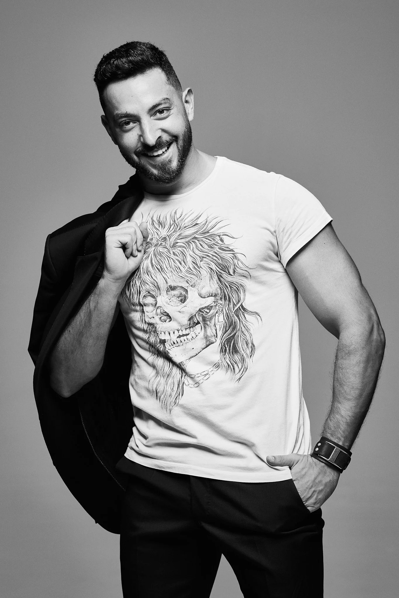 Portrait photographer in Dubai. A smiling man with a beard, wearing a graphic t-shirt with a skull and lion design, holding a jacket over his shoulder against a plain background.
