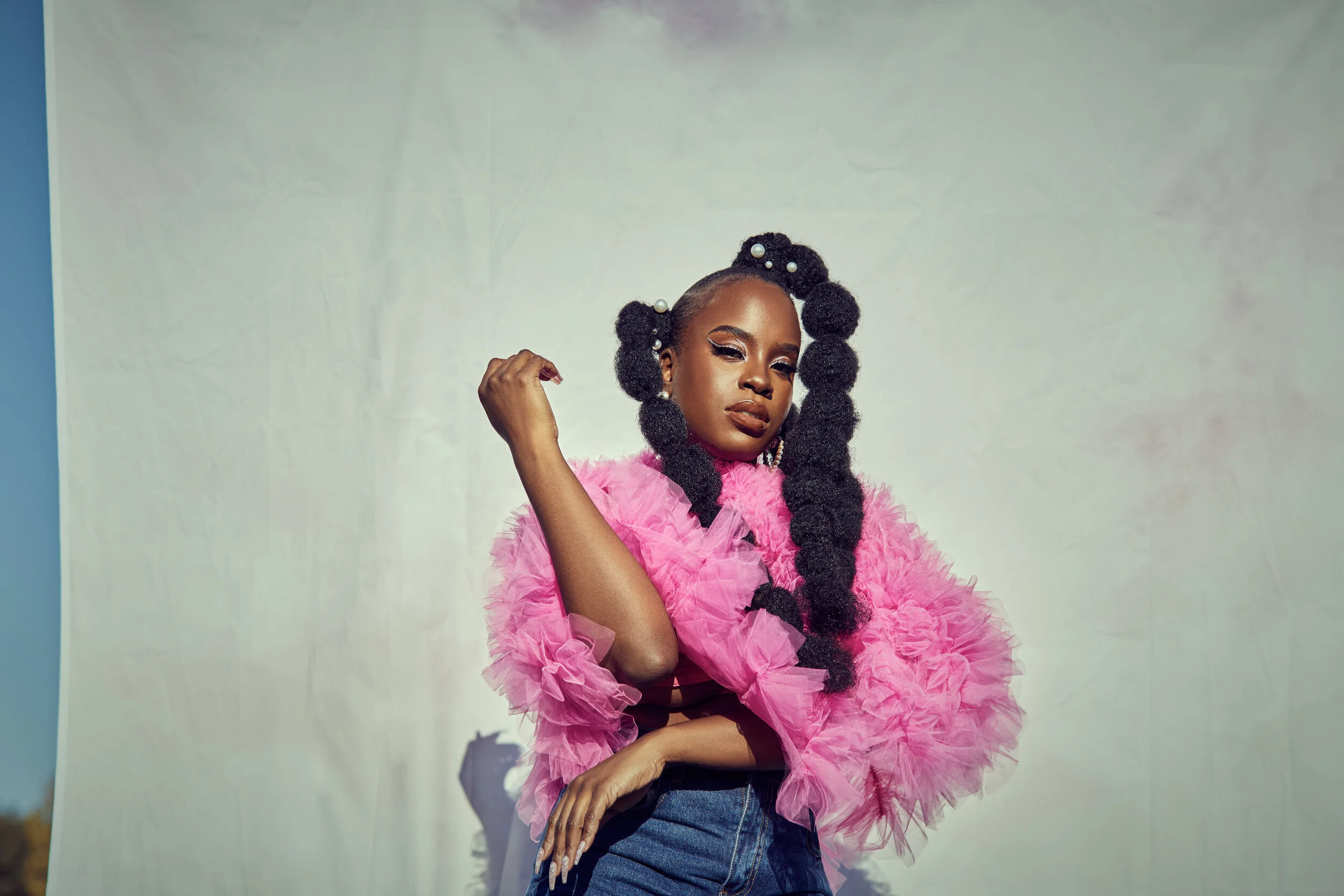 A woman posing in front of a neutral background, wearing a vibrant pink ruffled top and high-waisted jeans, with elaborate black braided hair decorated with white pearls, and bold makeup with dramatic eyeshadow and lipstick.