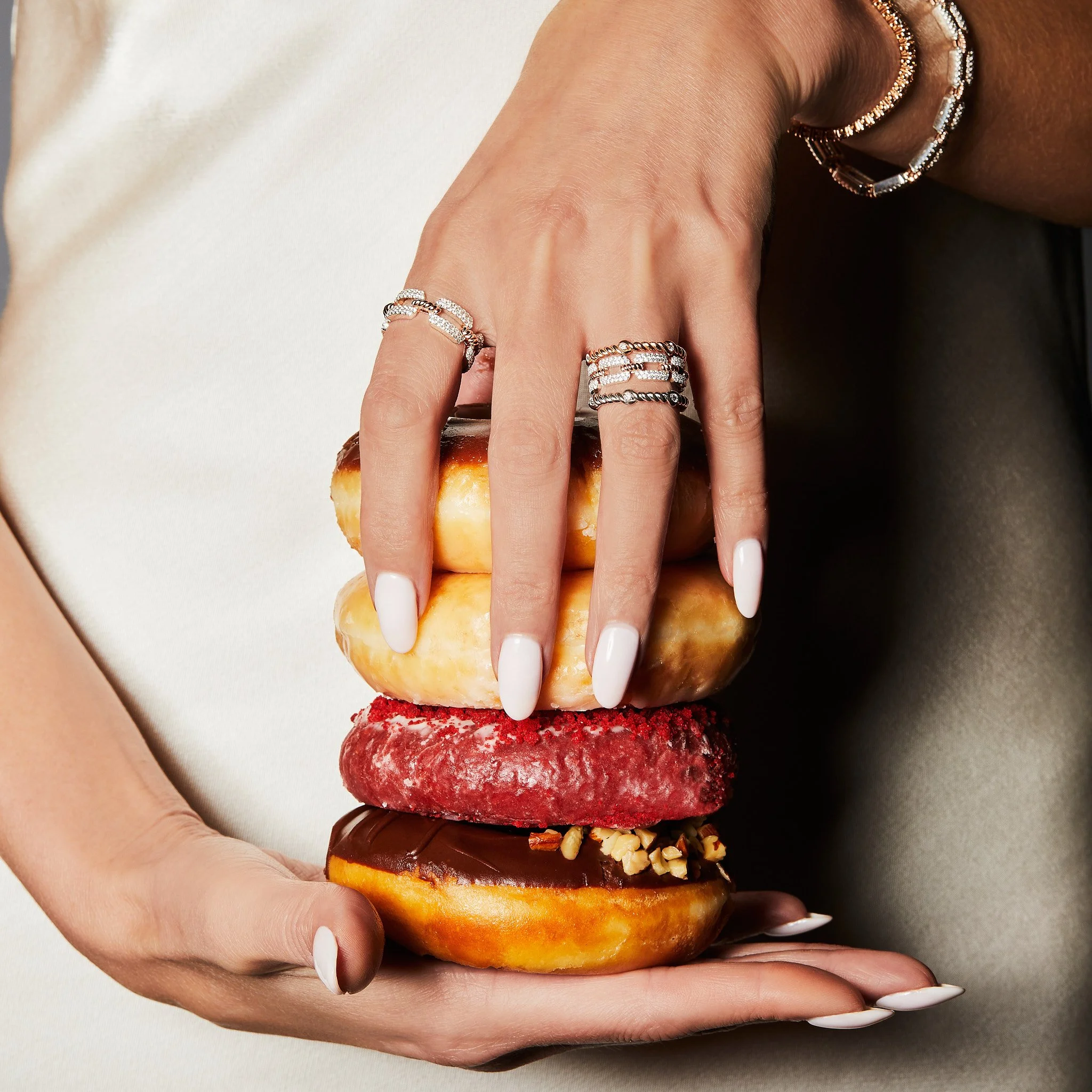 A hand with manicured white nails and jewelry, holding a stack of four donuts with different toppings and glazes.