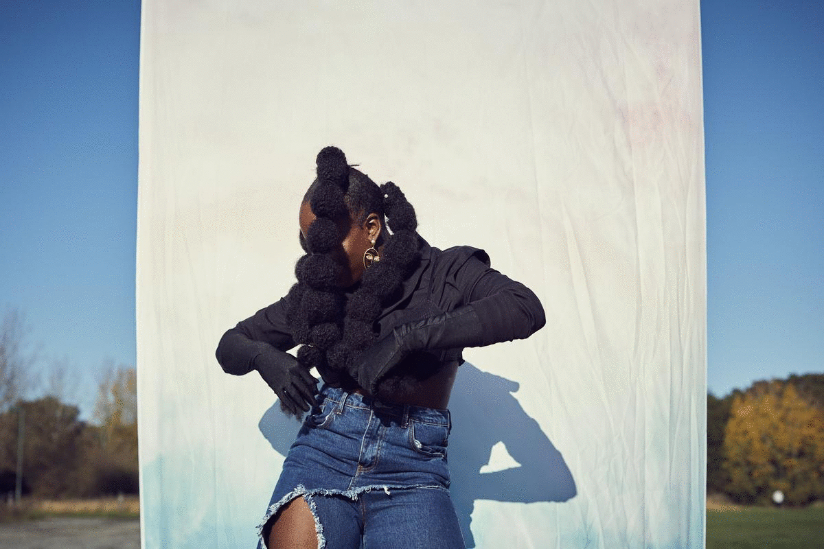 A woman with braided hair styled in large loops stands outdoors in front of a white backdrop during daytime. She is wearing a black long-sleeve top, ripped denim shorts, black gloves, and gold hoop earrings. Her face is partially obscured as she adju