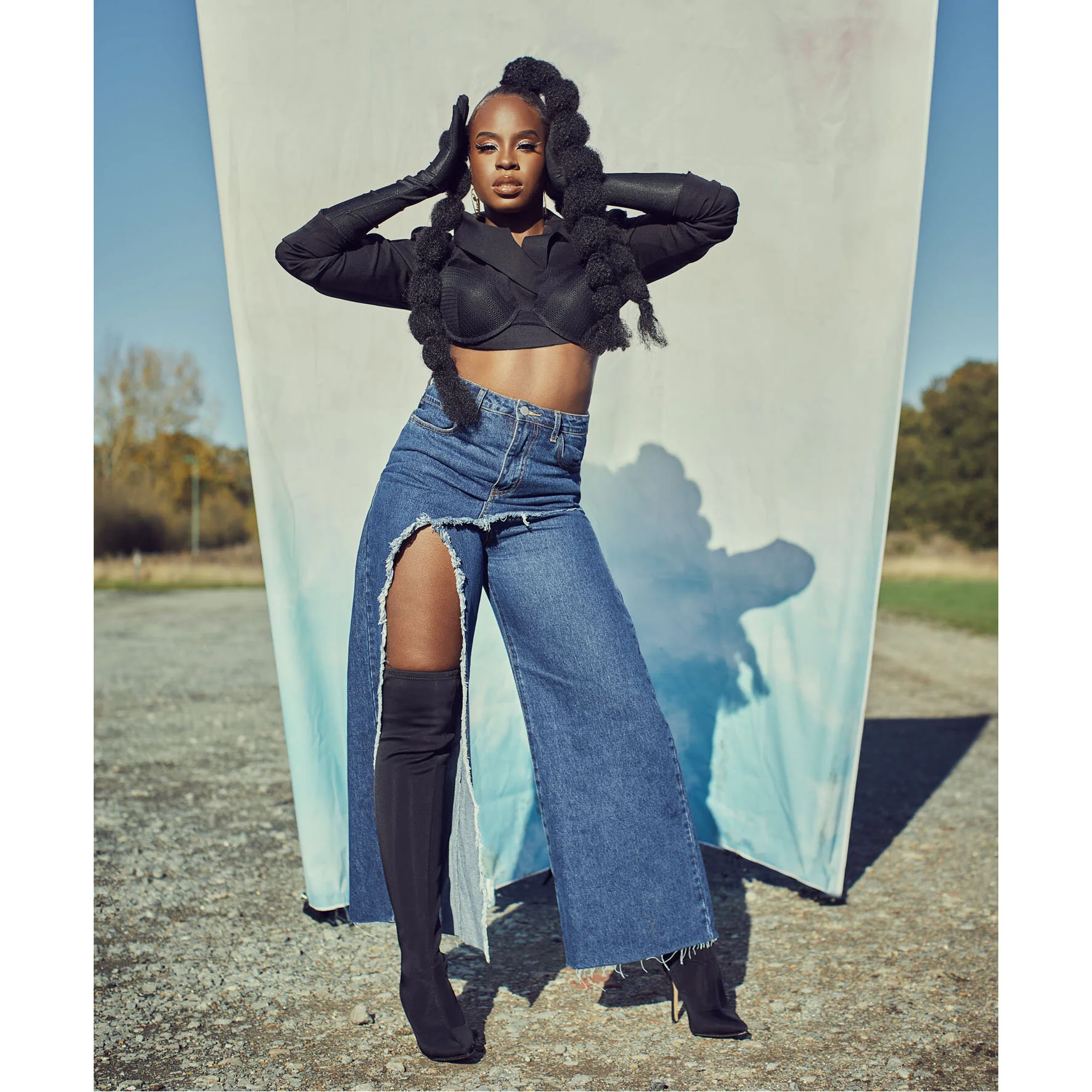 A woman with long, braided hair and dark makeup standing outdoors in front of a light-colored backdrop, wearing a cropped black top, ripped wide-leg jeans with a thigh-high slit, black over-the-knee boots, and black gloves, striking a pose with hands