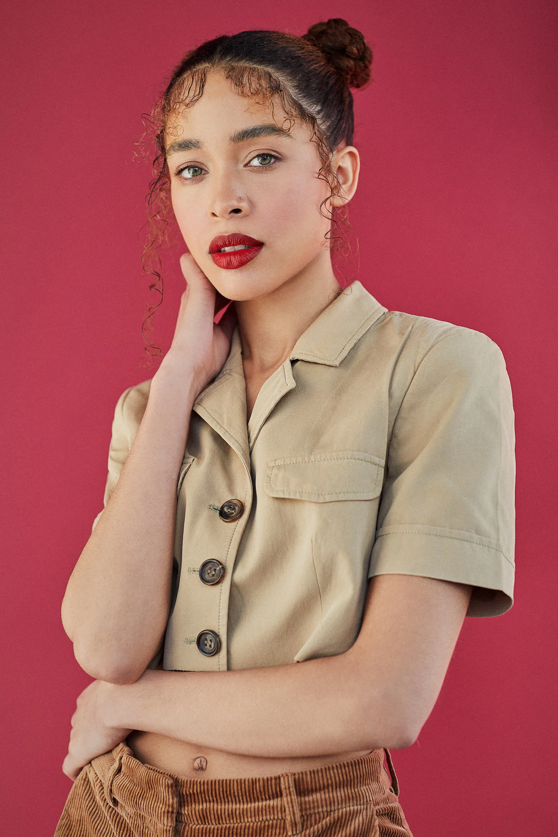 Portrait of a young woman with curly hair tied in a bun, wearing a beige button-up shirt and brown pants, posed against a red background.