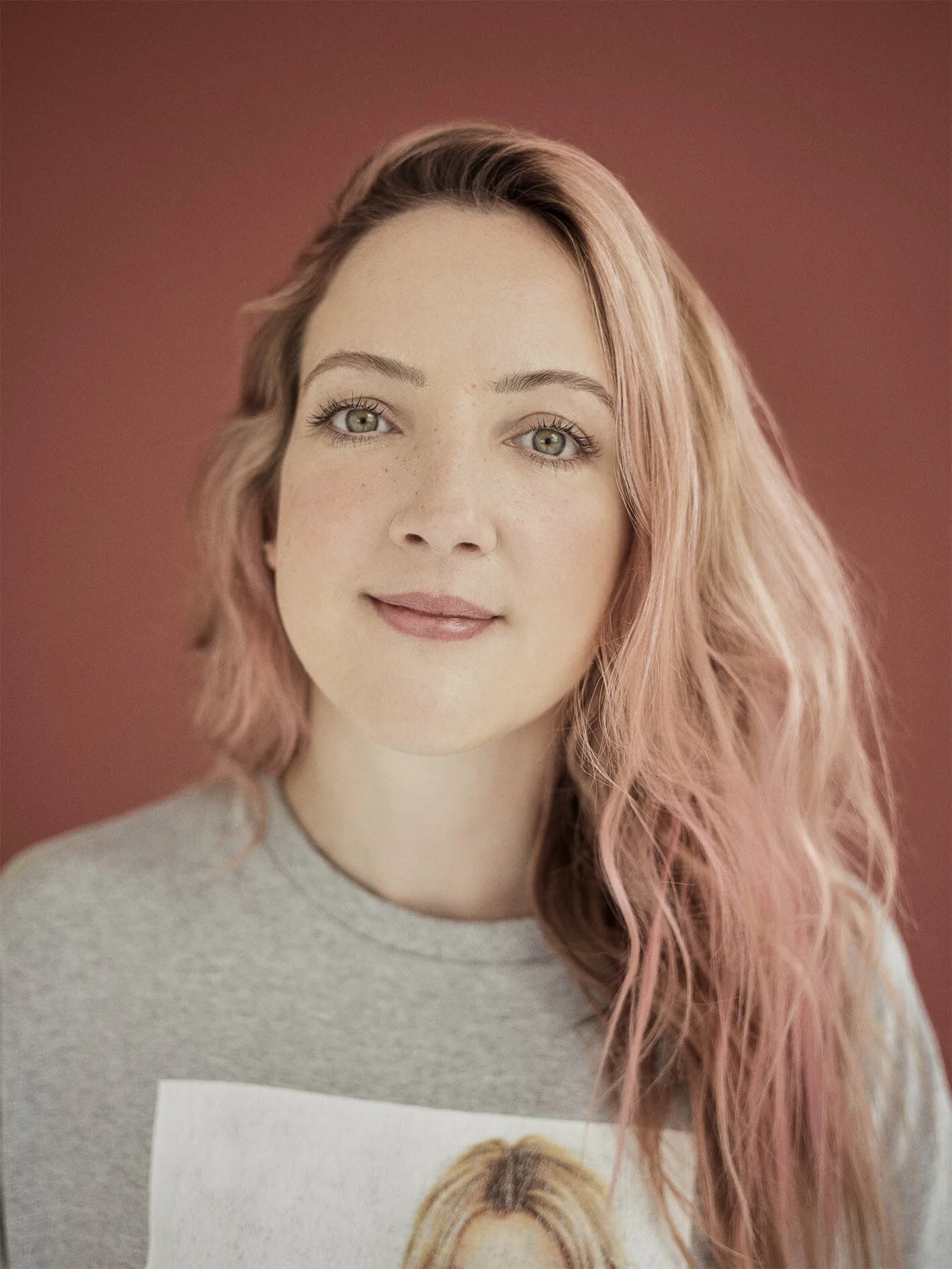 Portrait of a young woman with light skin, pinkish-blonde wavy hair, green eyes, and a soft smile, wearing a gray shirt with a graphic print, against a reddish-brown background.