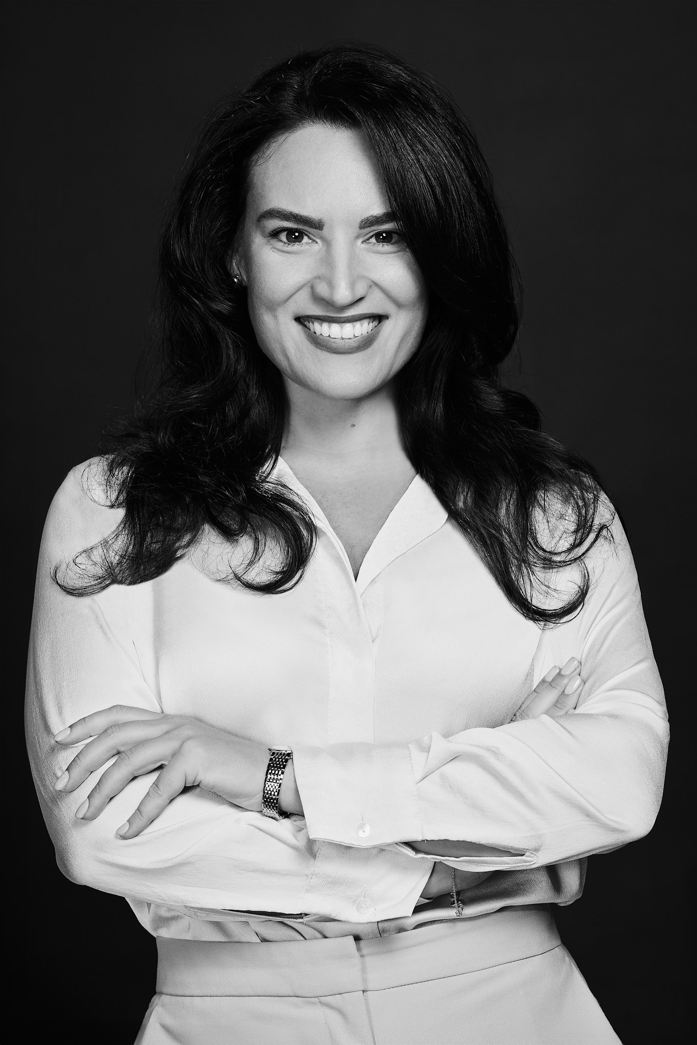 Portrait photographer in Dubai. Black and white portrait of a woman with shoulder-length dark hair, smiling, wearing a light-colored blouse with sleeves rolled up and a watch on her left wrist, against a dark background.