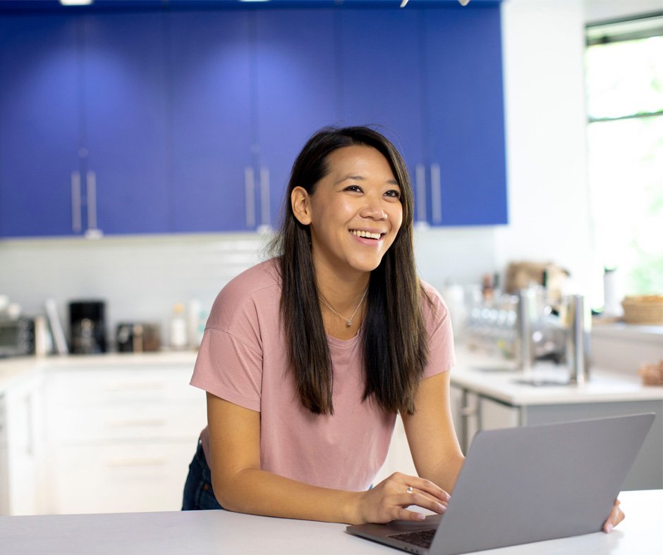 A young lady is smiling, she is standing at her kitchen bench, she is using her laptop to research what the Australian Taxation Office will be targeting in tax return lodgements.