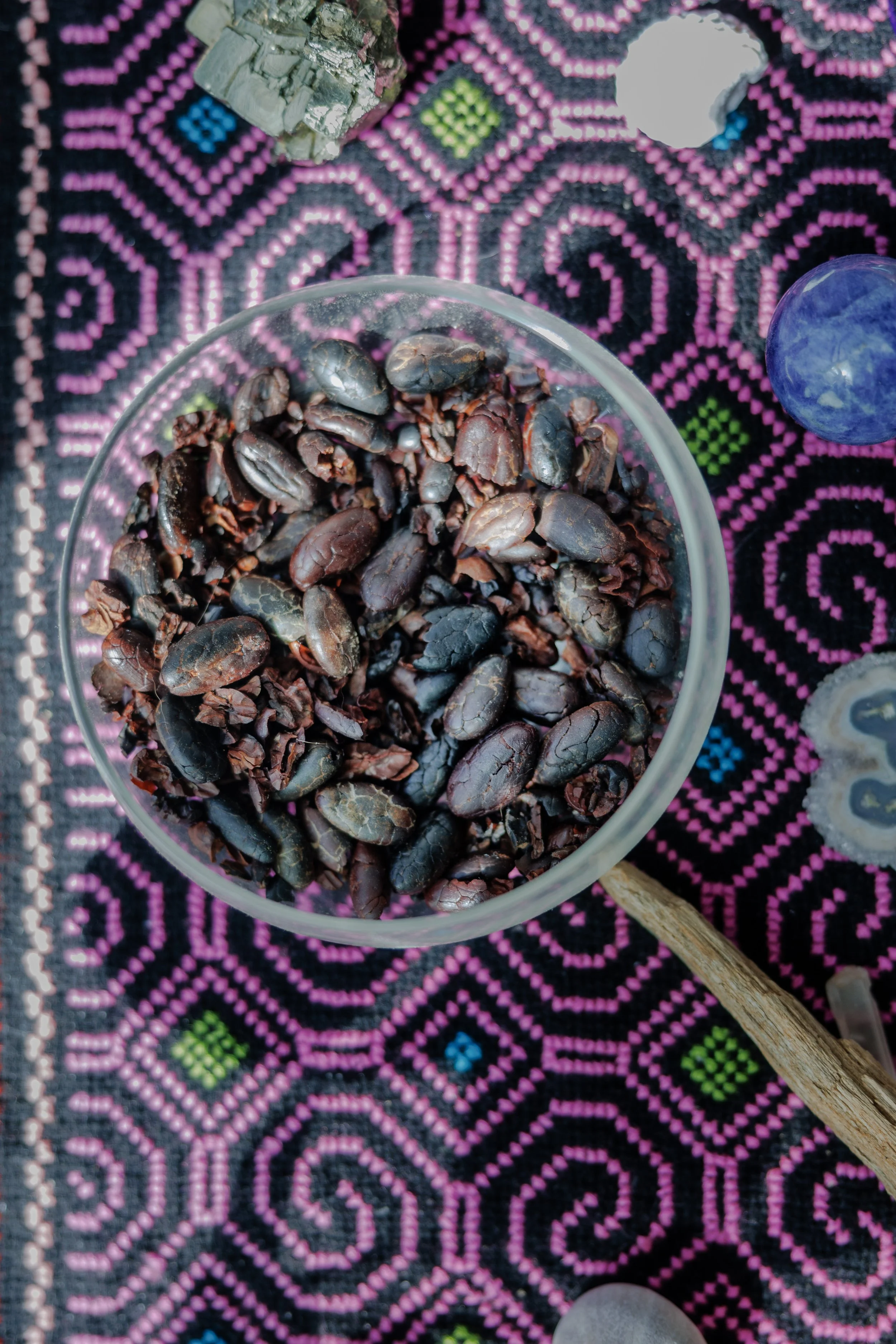 A small glass bowl filled with cacao beans on a colorful, patterned Shipibo textile with geometric shapes in pink, black, green, and blue.