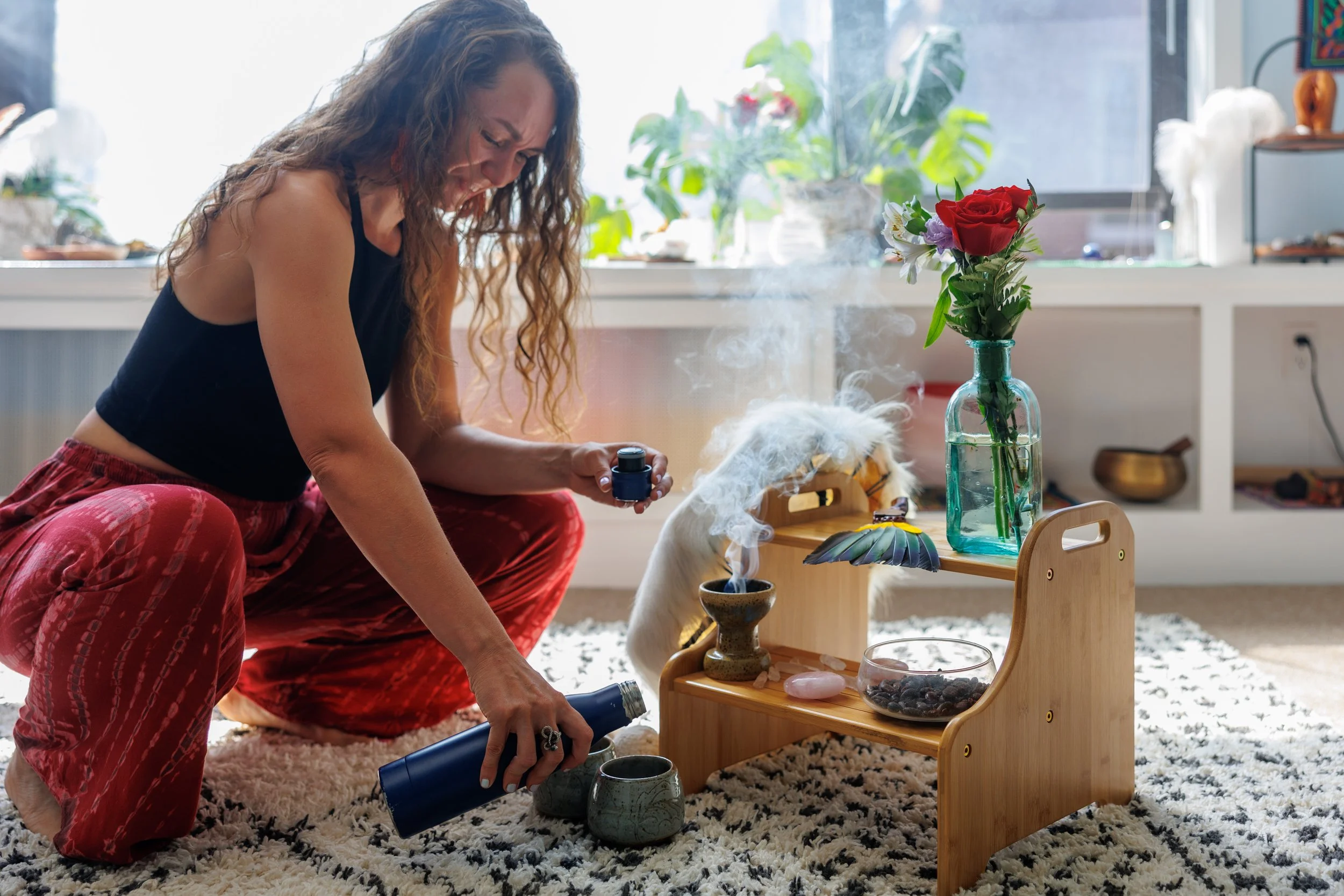 Ariel Szabo, a sacred sexuality and  plant medicine practitioner, is performing a smoke aromatherapy ritual. She is kneeling beside a wooden table with various objects, including a vase with roses and other flowers, pouring cacao for a ceremony