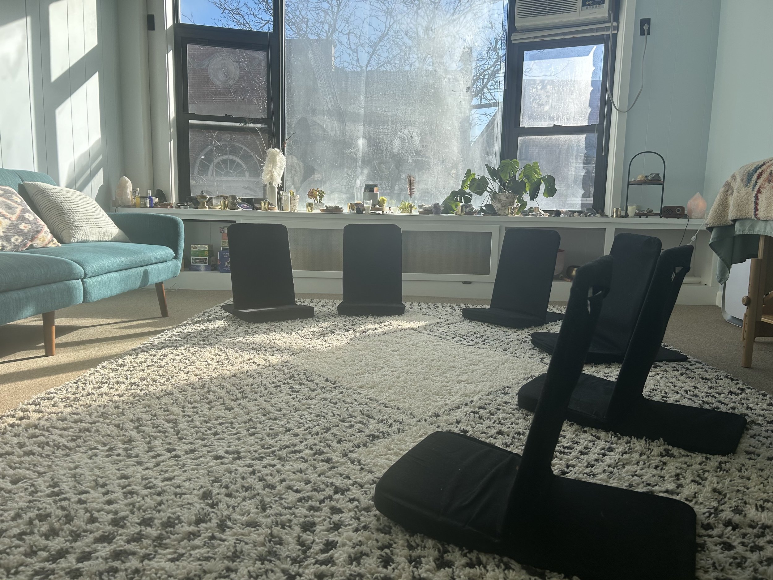 Sunlit living room with a teal sofa, black meditation chairs, a white shag rug, and large windows with a view of trees outside.