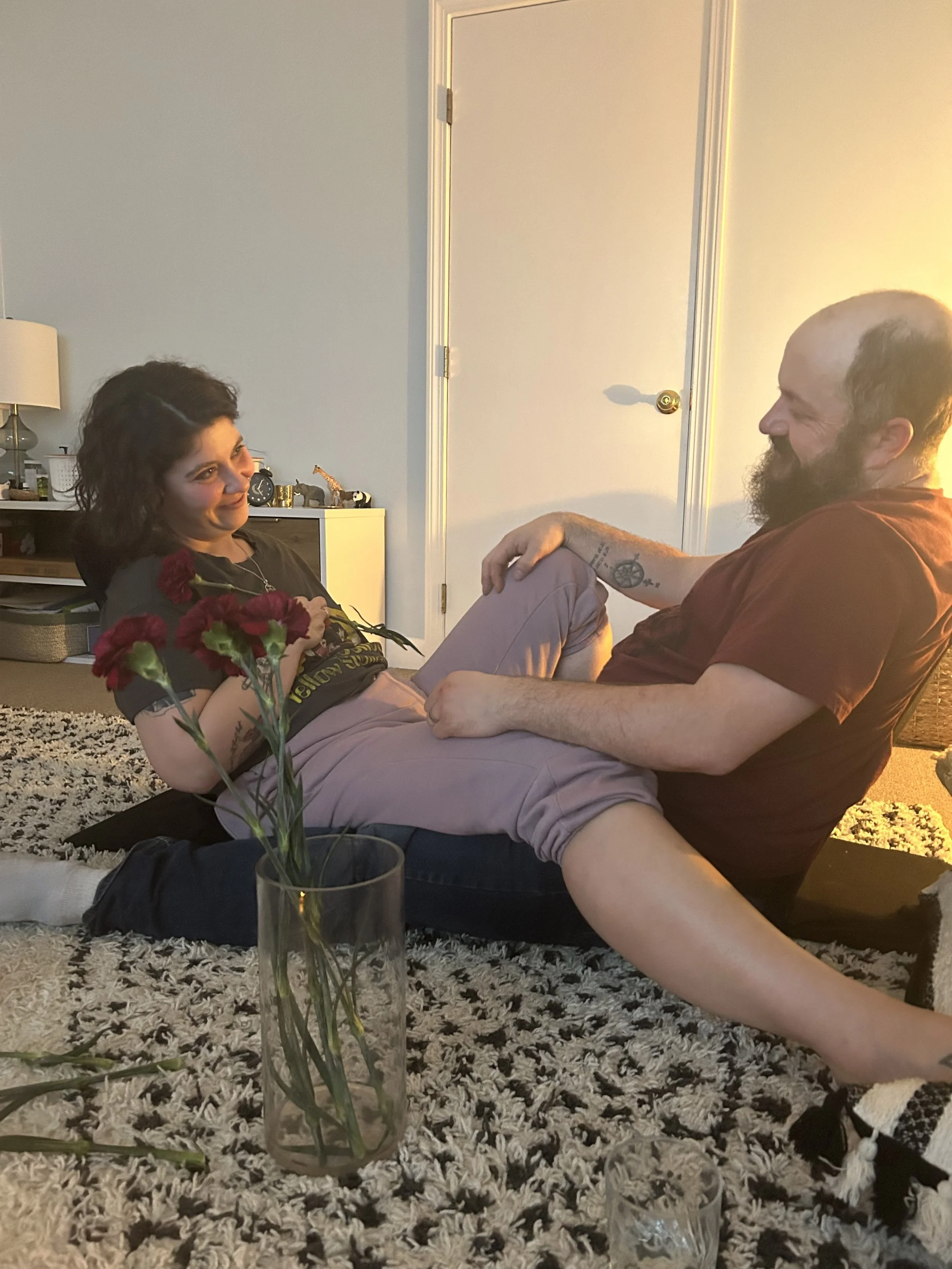 A man with a beard and tattoos on his arm sitting on the floor, holding a woman's leg. The woman is smiling, holding red flowers, and sitting on the floor. They are sharing an intimate moment in a couples cacao ceremony.