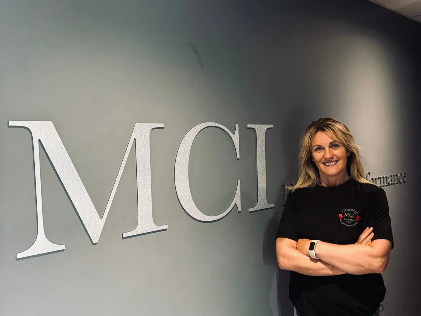 Woman with blonde hair smiling, standing with arms crossed next to an 'MCI' sign on a gray wall.