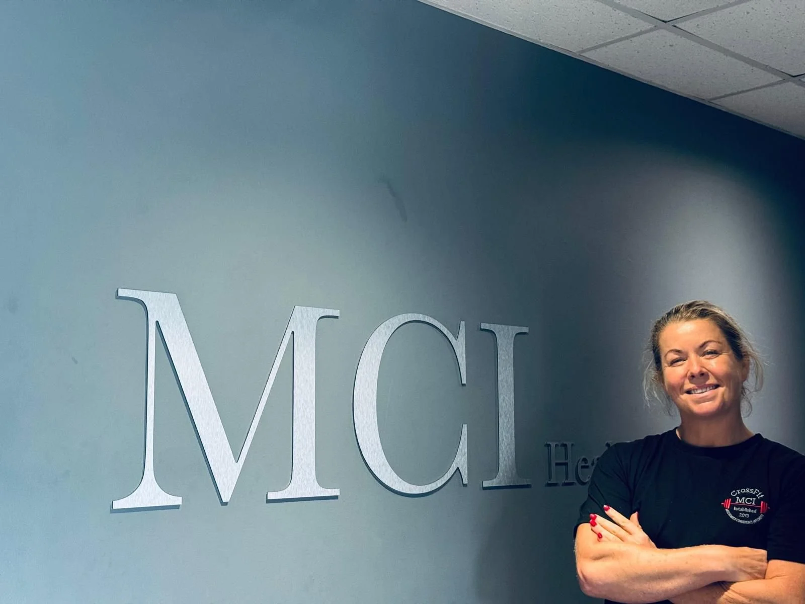 A woman stands smiling with her arms crossed in front of a wall with large letters spelling MC and part of the word 'Health'. She is wearing a black t-shirt with a logo on it.